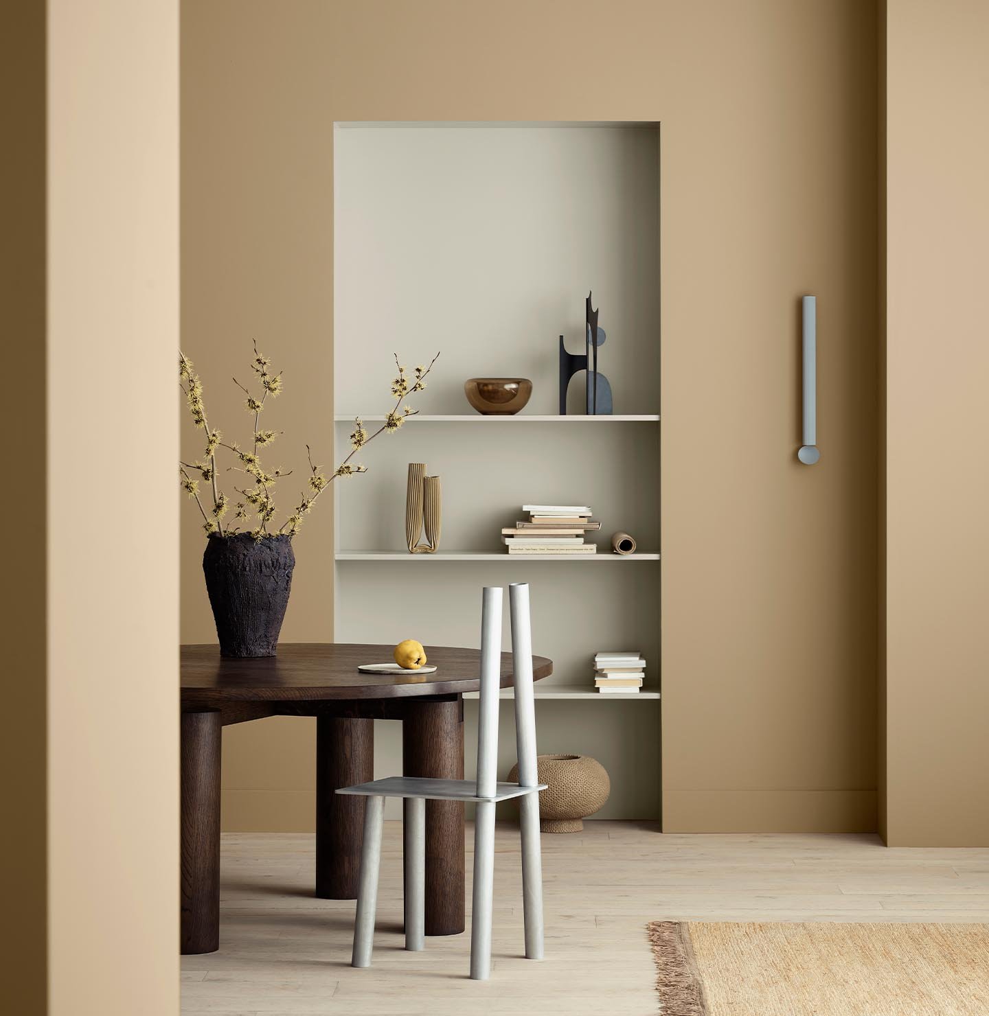 A warm, minimal dining space with golden brown walls painted with Jotun 1632 Rock Sugar, and built-in shelving, painted with Jotun 12075 Soothing Beige, styled with books and ceramics.