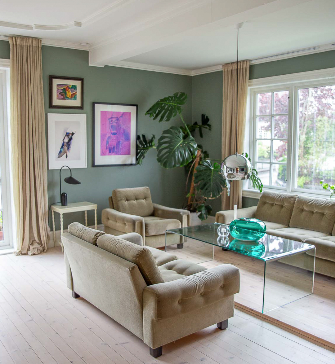 Stue med stor terrassedør og vindu med sprosser, lyst tregulb og vegger malt grønne i Jotun LADY Antique Green, tak med ornamenter malt hvitt i fargen Bomull fra Jotun. Grønnbeige salong, glassbord og beige gardiner.