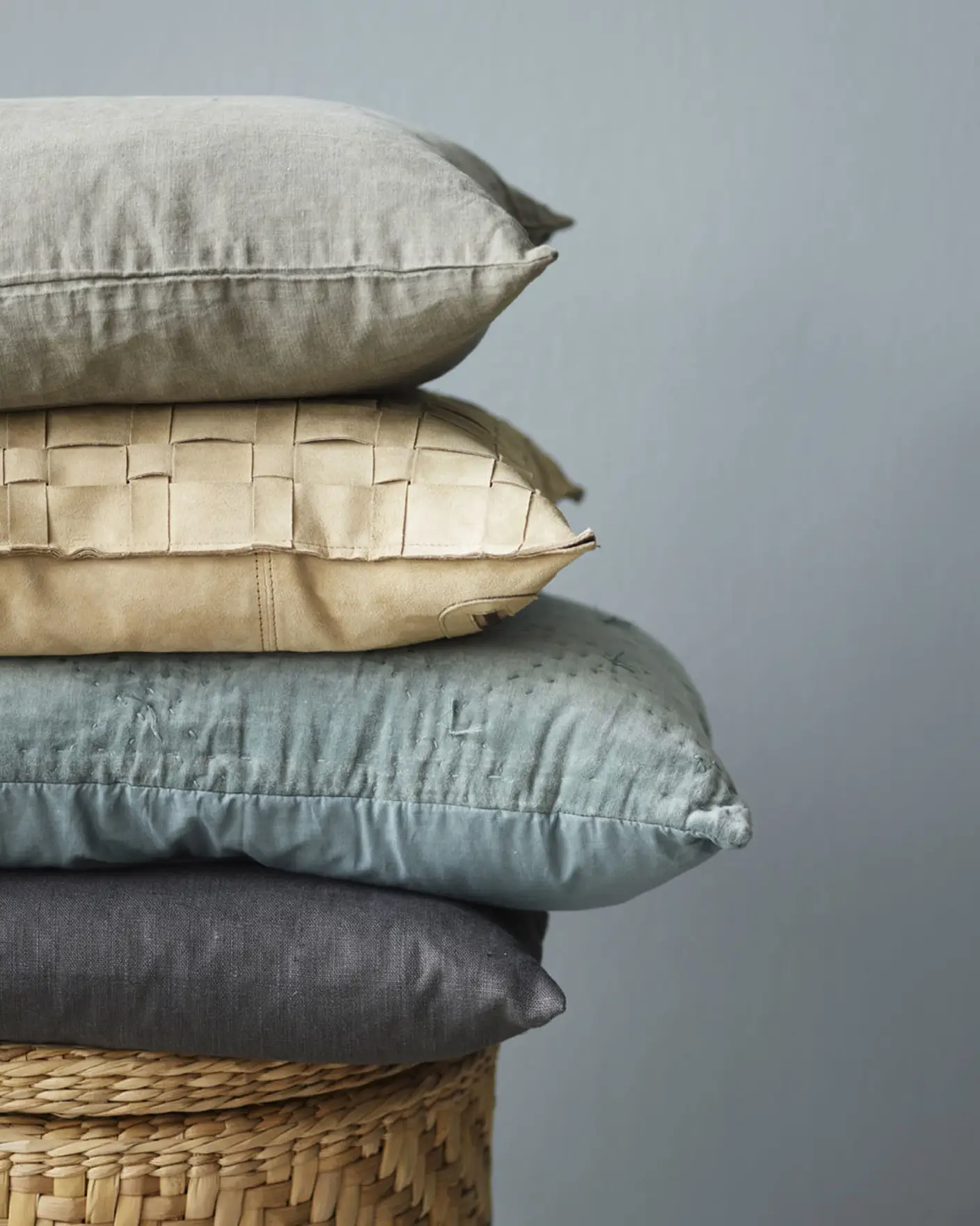 Stack of pillows in muted earthy and blue tones, resting on a woven basket with a muted, greenish blue wall in the background painted with Jotun 5249 Artic Grey.
