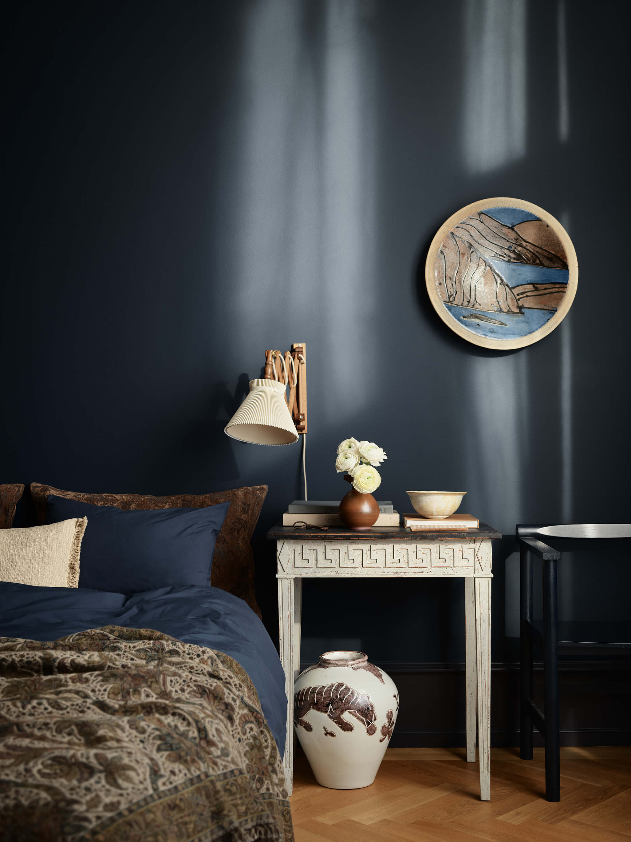 Et stemningsfullt soverom med dyp marineblå vegger i Jotun 4403 Midnight Blue, et vintagehvitt nattbord, en vegglampe og matchende blått sengetøy