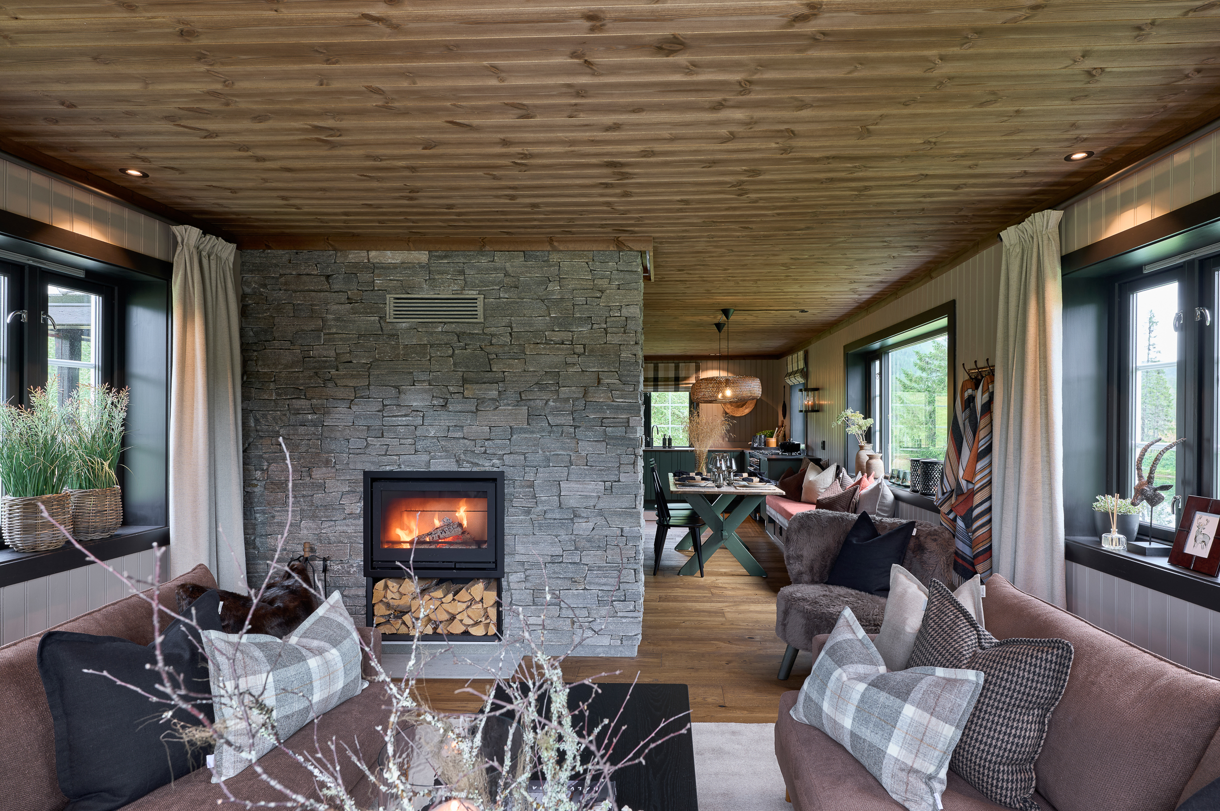 Hyttestue med stor skiferlagt peis, panel på vegger og tak, grønt kjøkken i enden.