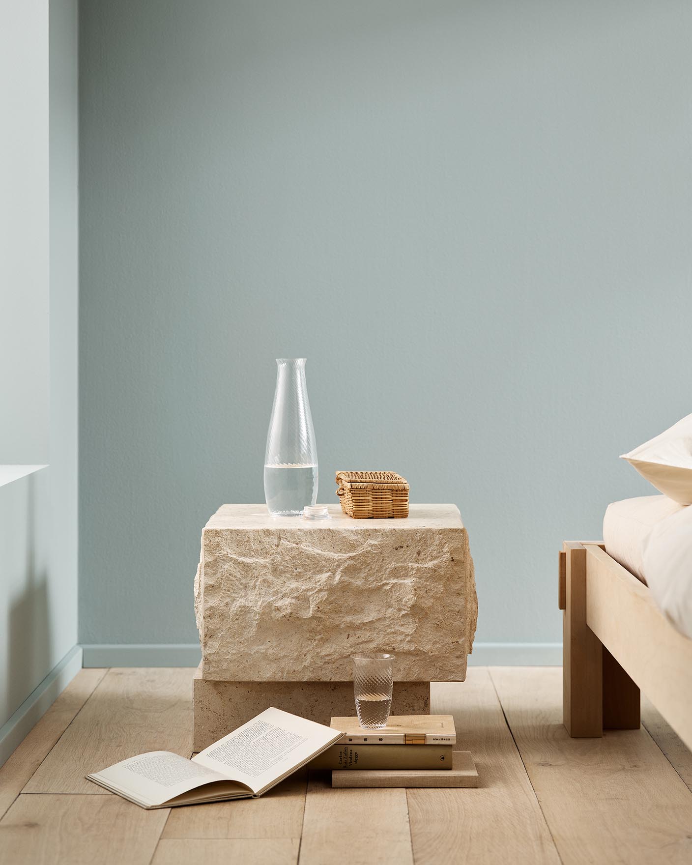 Minimalist bedroom with a stone bedside table and books on a wooden floor against a muted blue wall painted with 6378 Iconic from Jotun.
