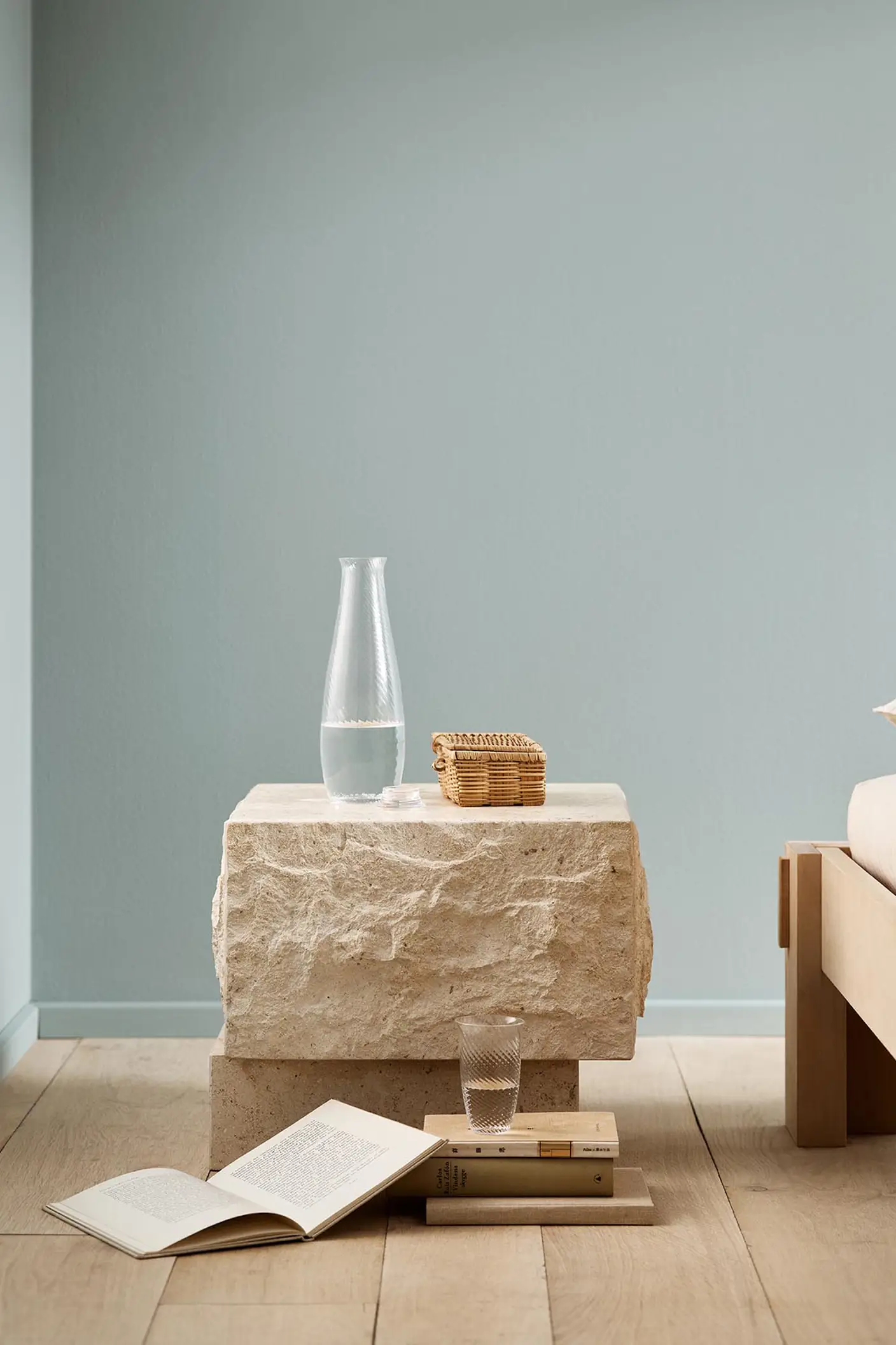 Minimalist bedroom with a stone bedside table and books on a wooden floor against a muted blue wall painted with 6378 Iconic from Jotun.