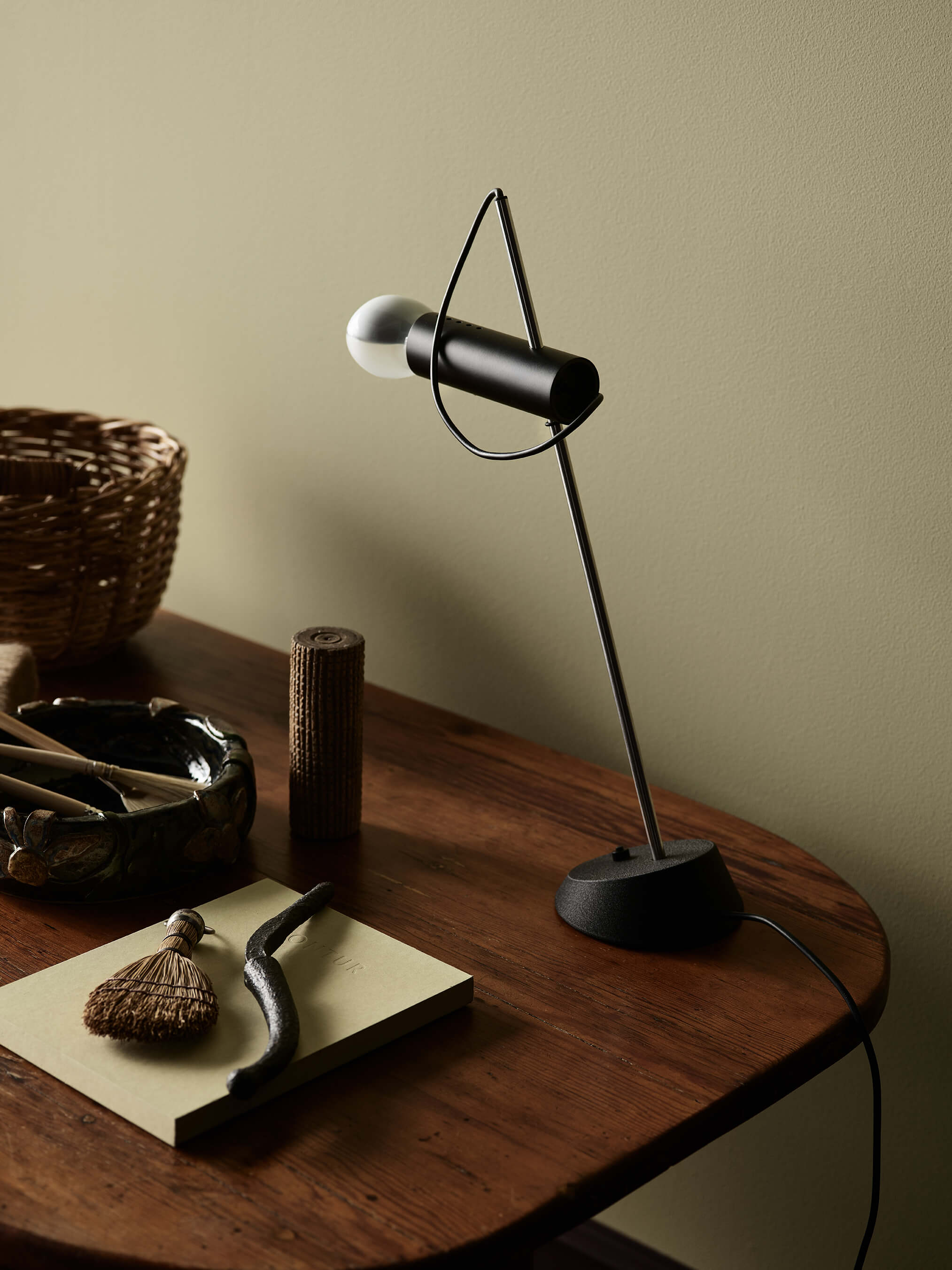 Et minimalistisk hjemmekontor med olivengrønne vegger i Jotun 0290 Biri, med en elegant svart skrivebordslampe på et bord i mørkt tre med dekorative gjenstander