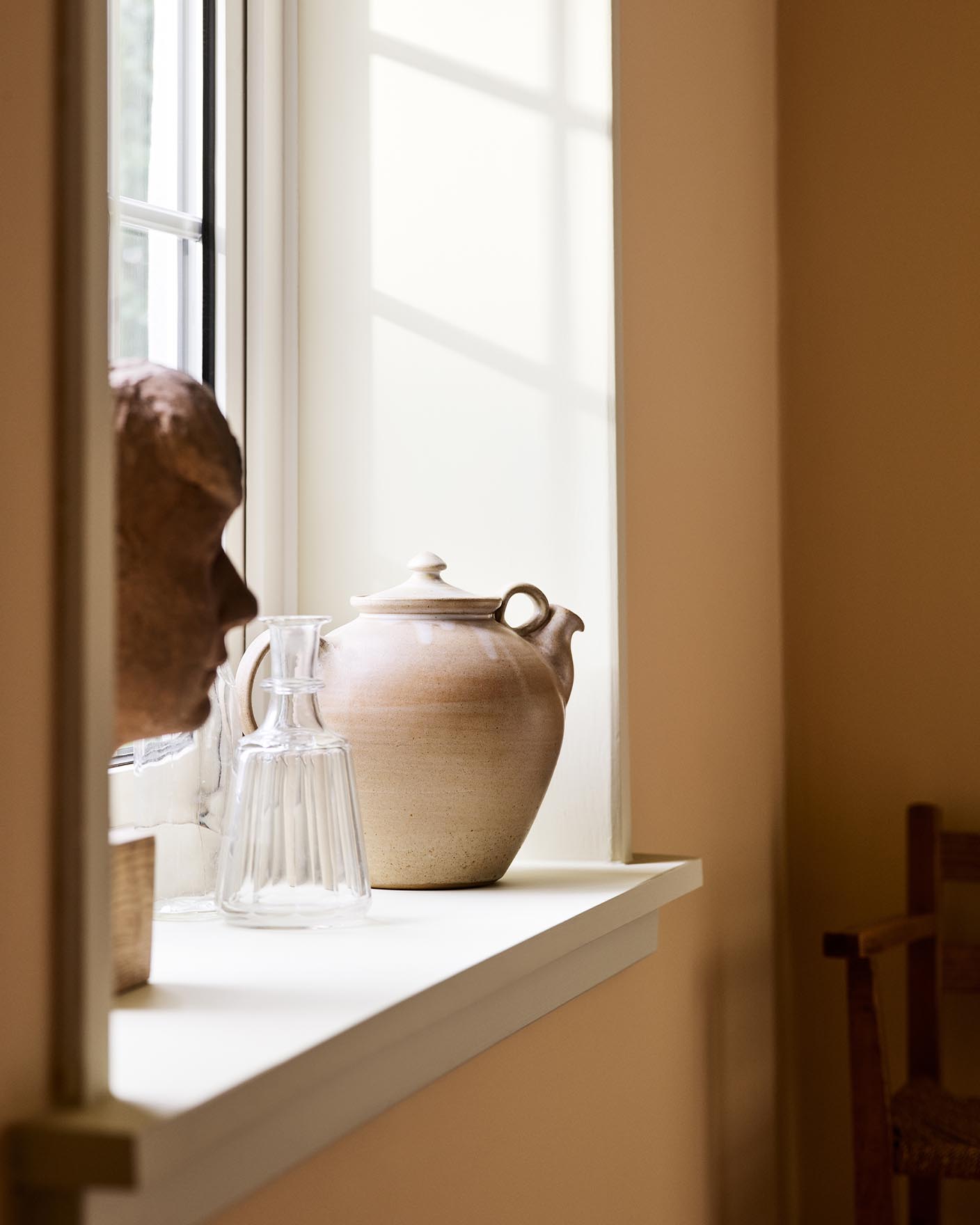 Ceramic teapot and glass pitcher with sunlight against a ochre yellow wall painted in the Jotun colour 11202 Mild Ochre.