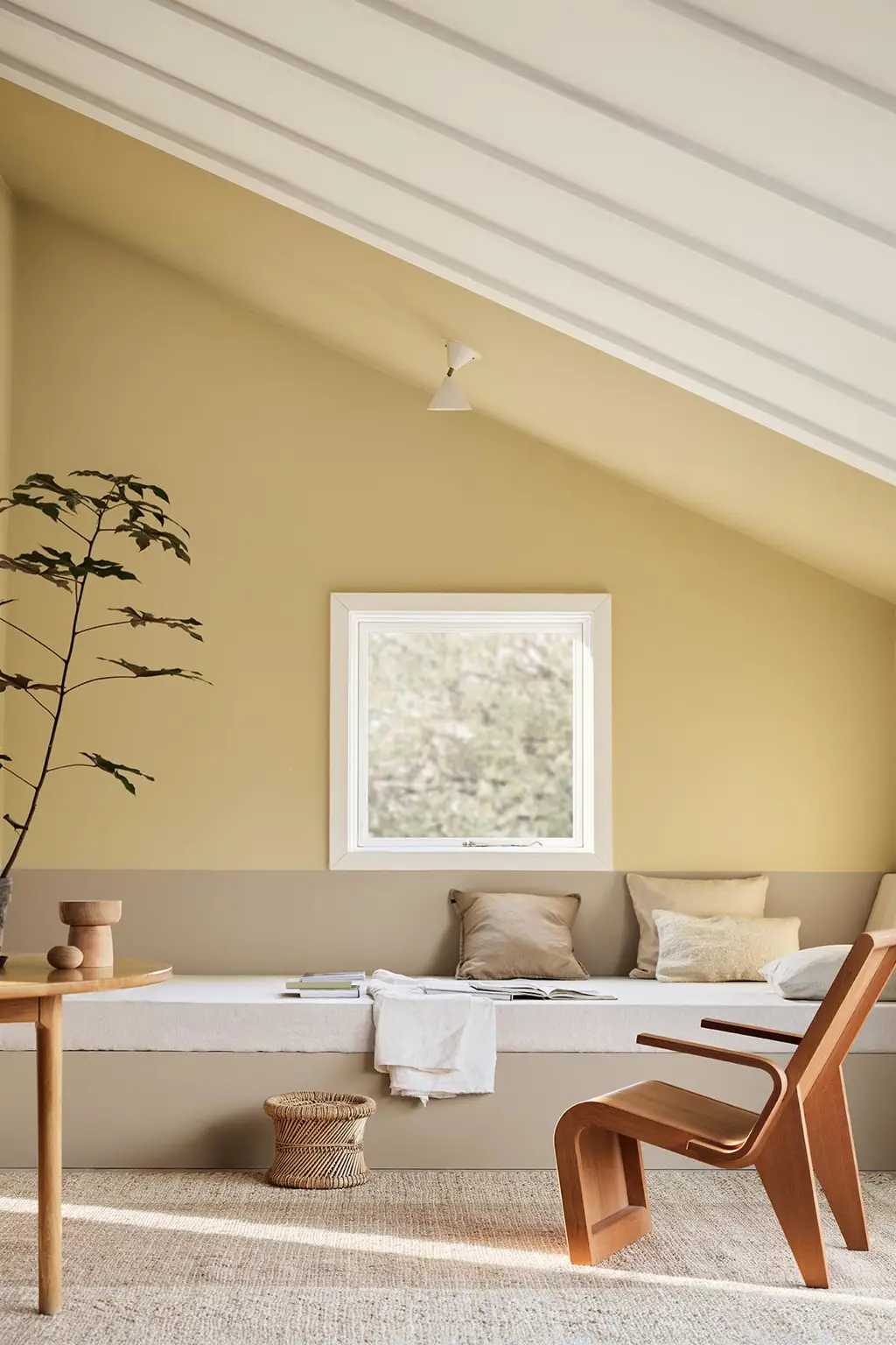 A daybed, wodden chair and a potted plant against muted, greenish yellow walls painted in the colour 1965 Ginger Tea from Jotun. Daybed painted in the colour 12300 Hazelnut Beige.