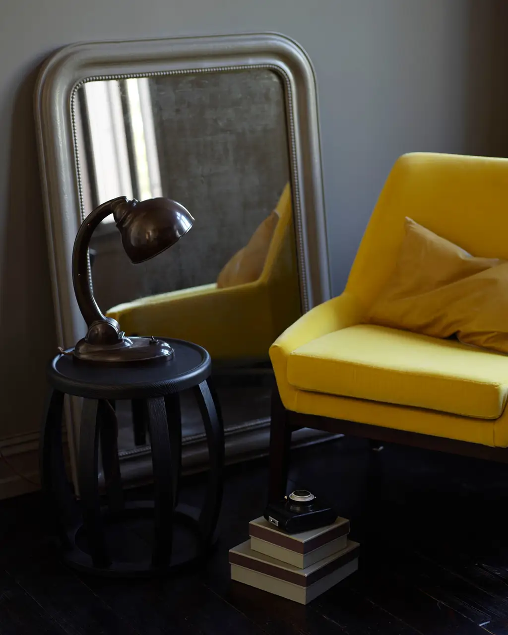 Silla amarillo mostaza, espejo vintage, mesita de madera oscura con lámpara industrial y cajas apiladas, todo frente a una pared gris marrón pintada con Jotun 2732 Deep Mocca.