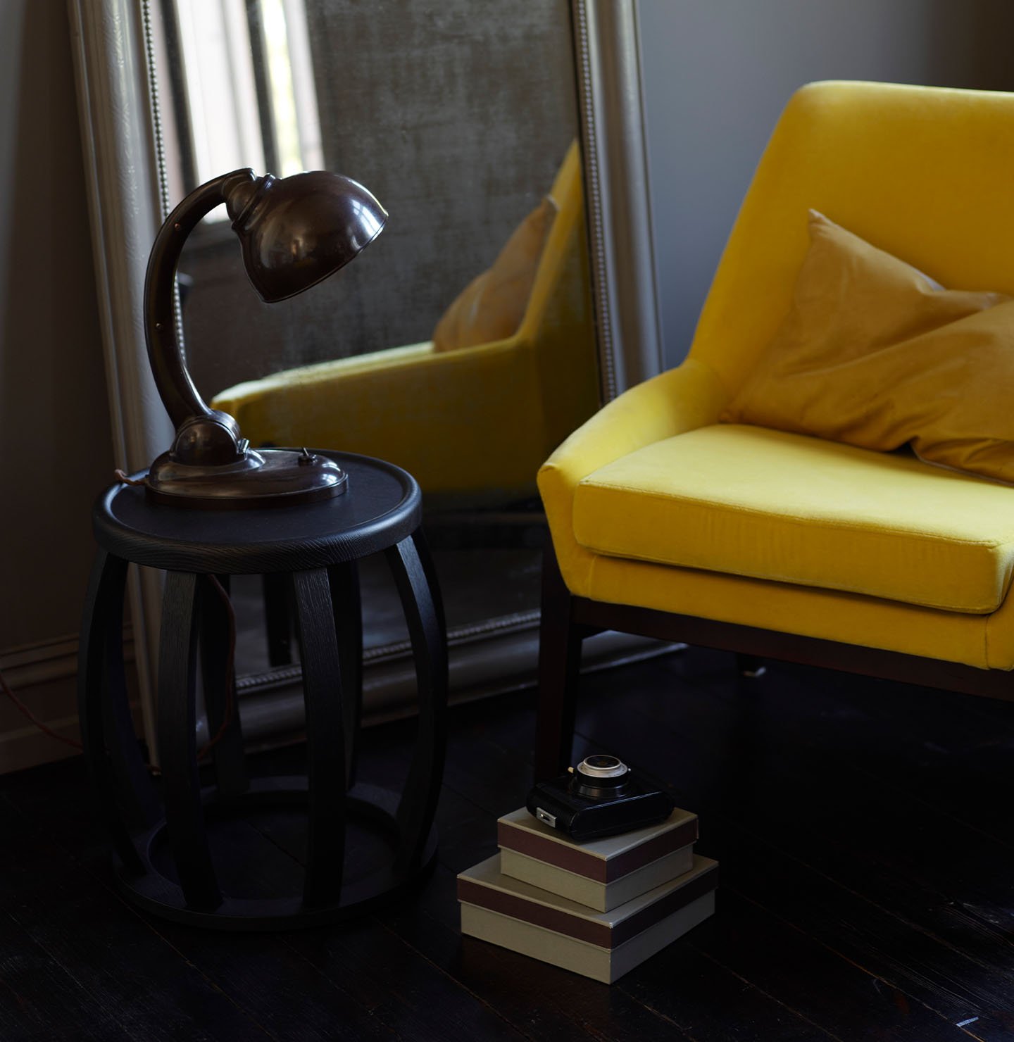 Mustard yellow chair, vintage mirror, dark wood side table with an industrial lamp and stacked boxes, all set against a brownish grey wall painted with Jotun 2732 Deep Mocca.