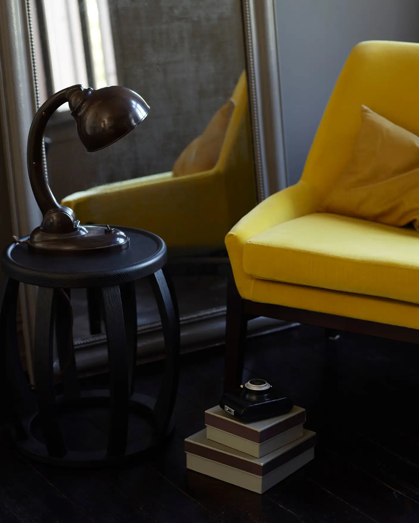 Mustard yellow chair, vintage mirror, dark wood side table with an industrial lamp and stacked boxes, all set against a brownish grey wall painted with Jotun 2732 Deep Mocca.