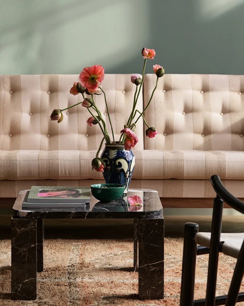 A dark marble coffee table holding a patterned blue vase with pink poppies, a small green bowl, and books, set against a tufted beige sofa and soft sage green wall
