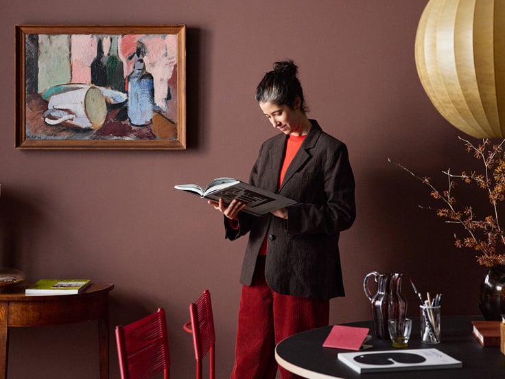 Person står og læser i en åben bog i et stilfuldt rum med væg malt i Sophisticated Red, moderne kunst på væggen og dekorativ lampe.