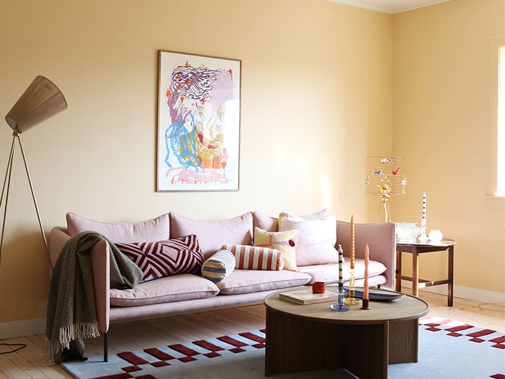Varmgul stue i Jotun Cheerful Peach med moderne rosa sofa, fargerikt bilde på vegg og mønstret teppe i rødt og lyseblått.