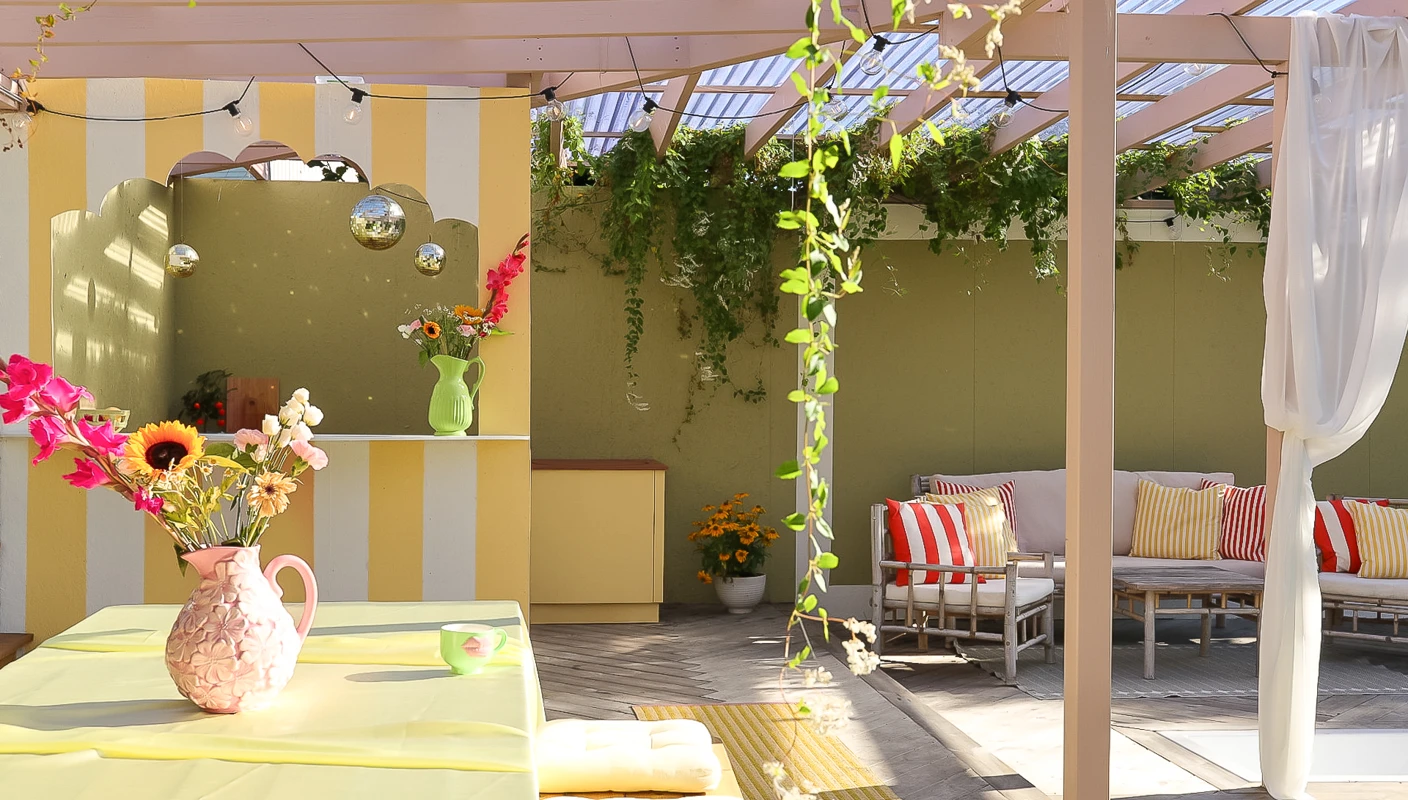 Utekjøkken på terrasse med rosa tak og stolper malt i Jotun Mellomrosa, med bar malt i gul og hvitstripet med Jotun Demigul og Jotun Bomull, grønn bakvegg i Jotun Grønnland, tropisk stemning med friske farger og blomster.