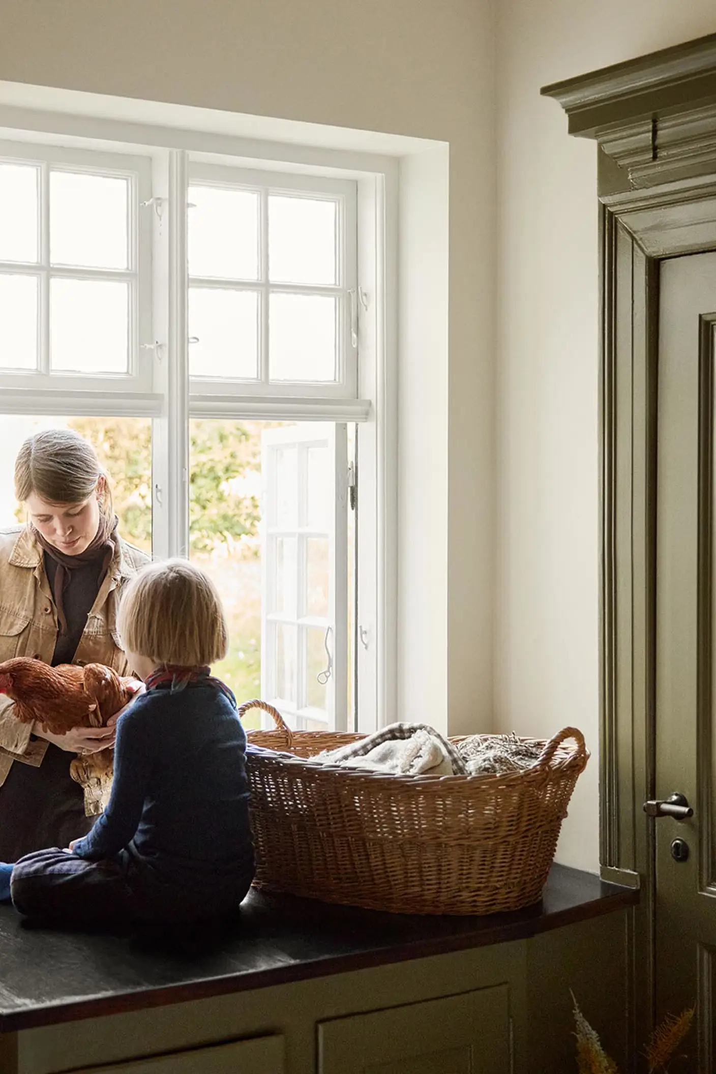 Woman and a child child on a kitchen counter by the window next to a olive-green cabinet in the colour 8597 Seaweed Green, against a muted yellow wall in the colour 12079 Gleam from Jotun.