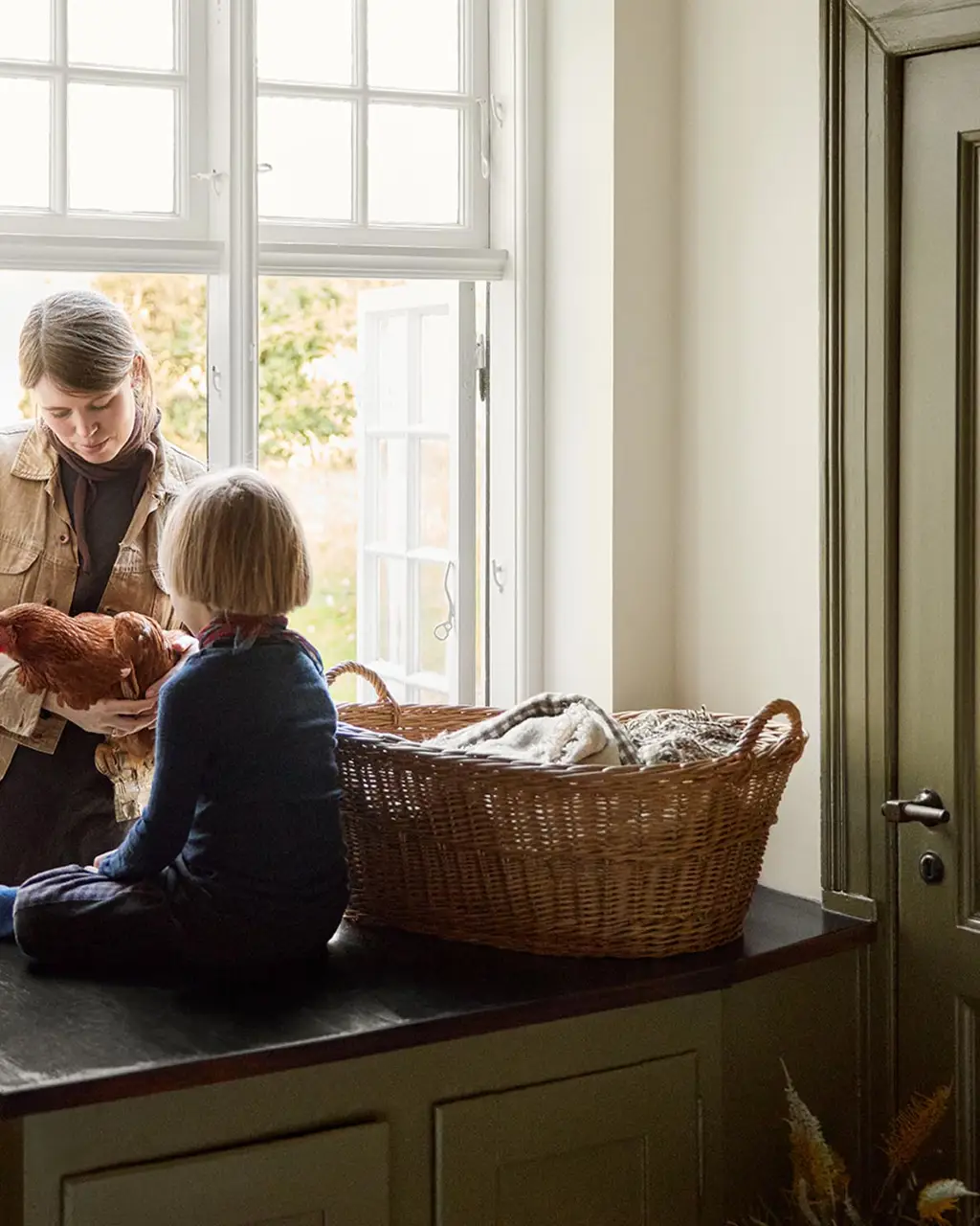 Woman and a child child on a kitchen counter by the window next to a olive-green cabinet in the colour 8597 Seaweed Green, against a muted yellow wall in the colour 12079 Gleam from Jotun.
