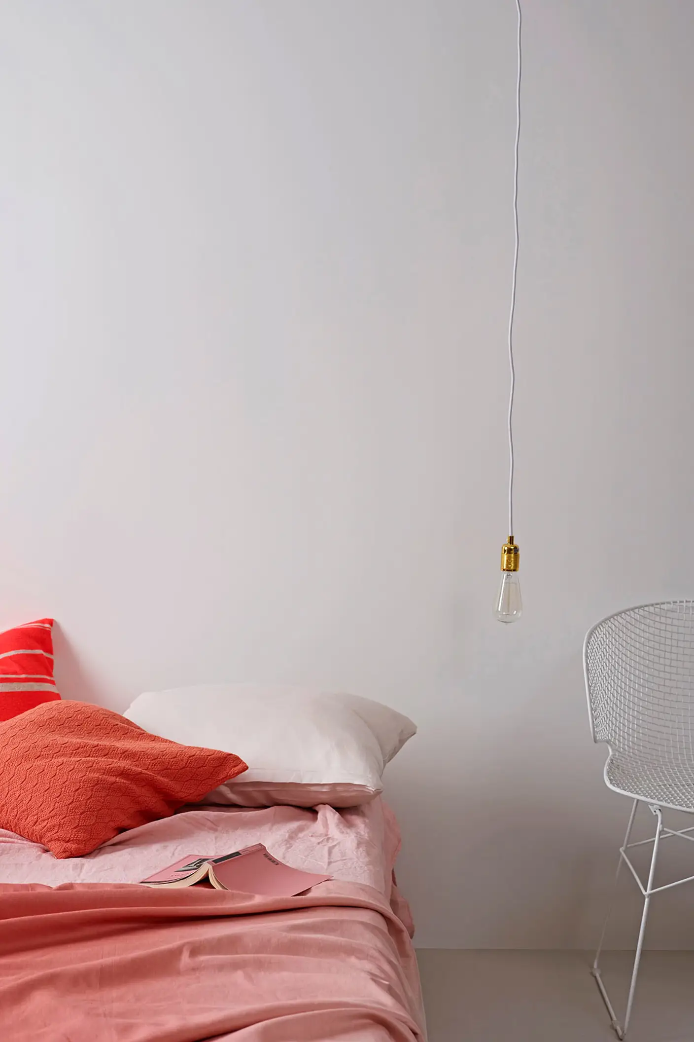 Mjukt rosa bäddset med korallfärgade accentkuddar på en låg säng, kombinerat med en vit trådstol mot en ljus, gyllene vit vägg målad med Jotun 1624 Lätthet.