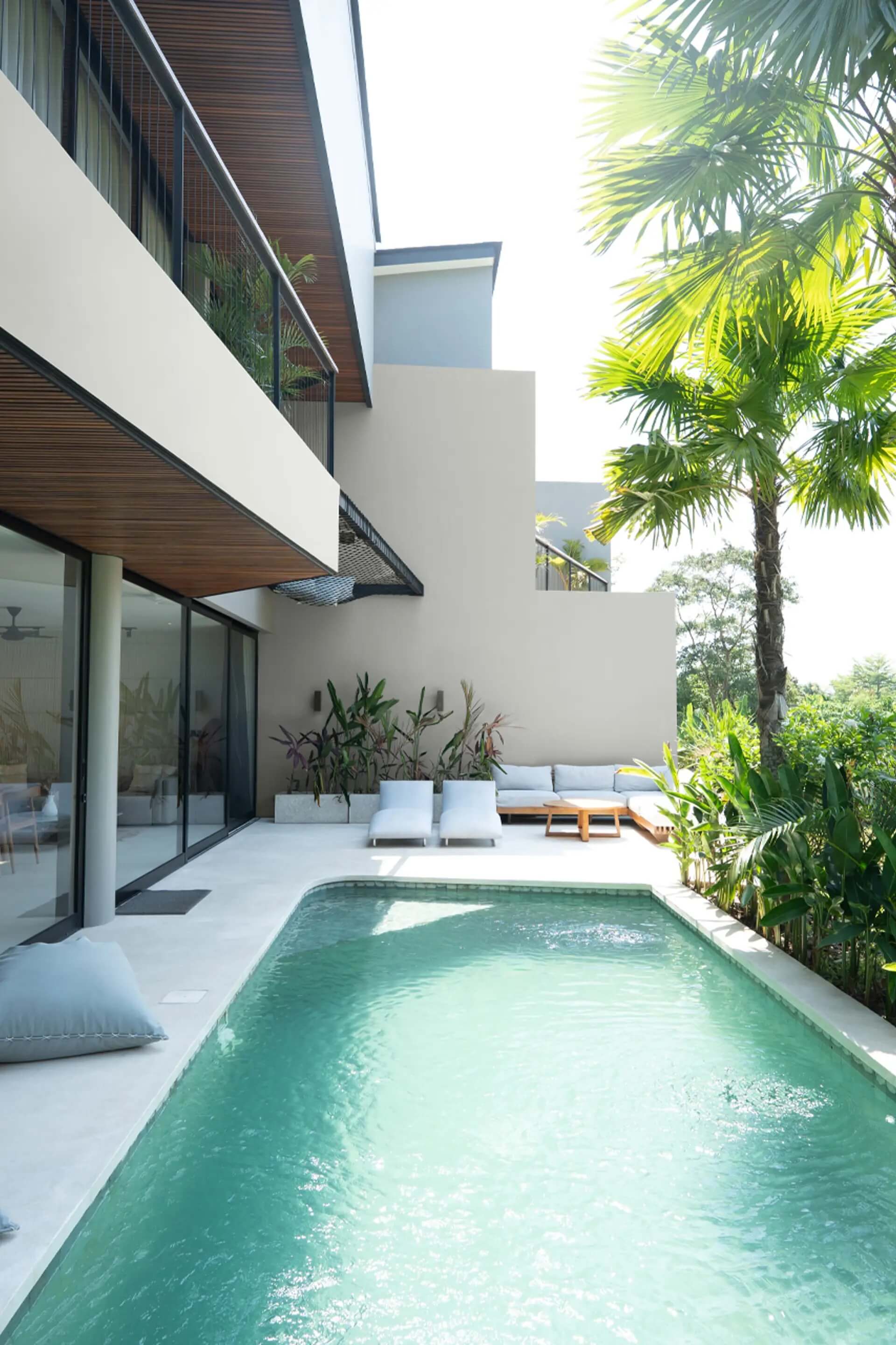 Modern luxury villa patio featuring a turquoise swimming pool, poolside lounge chairs, an outdoor sectional sofa, and lush tropical greenery
