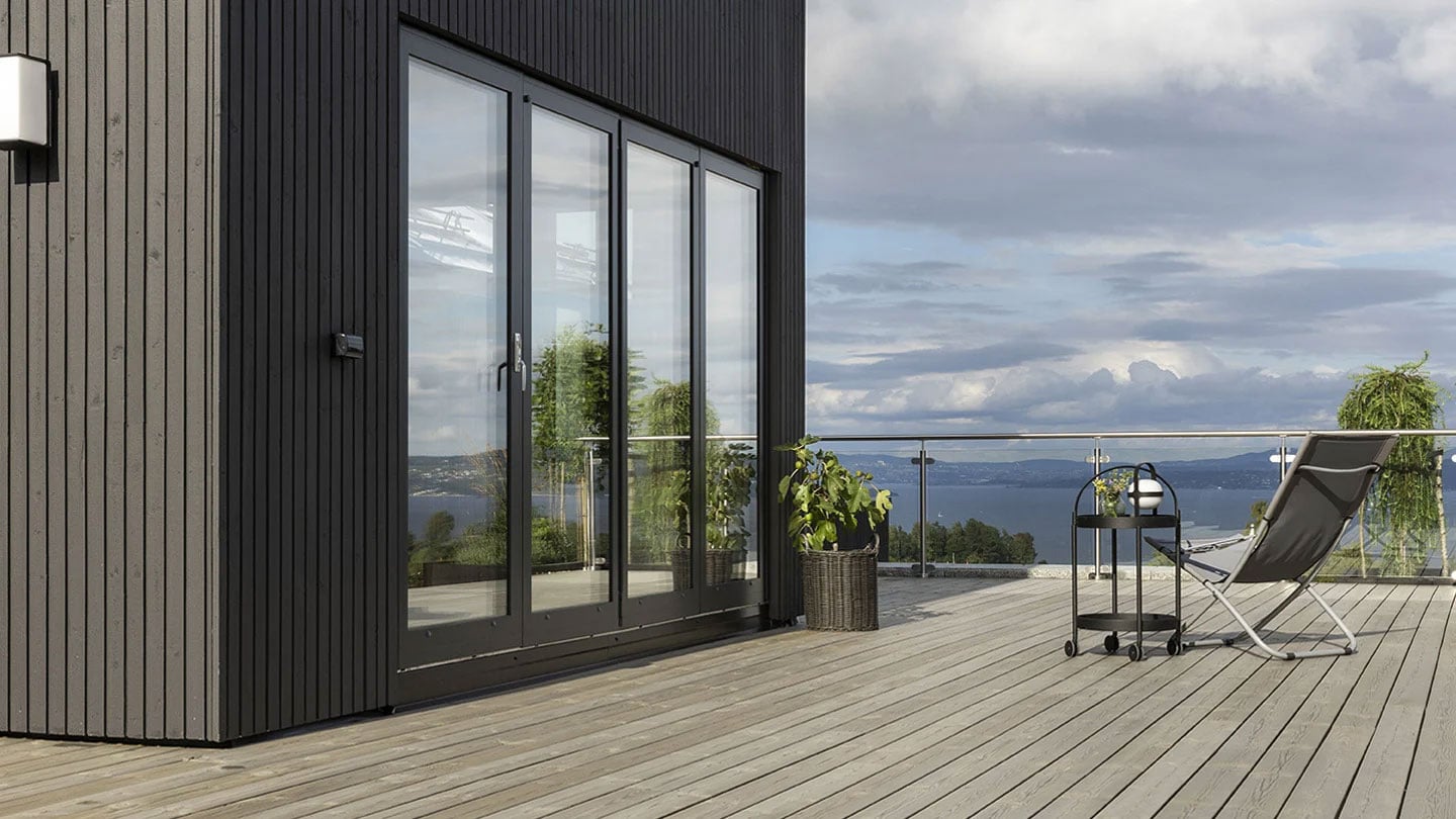 Moderne terrasse i træ med glasrækværk, loungestol og udsigt over vandet ved et sort træhus