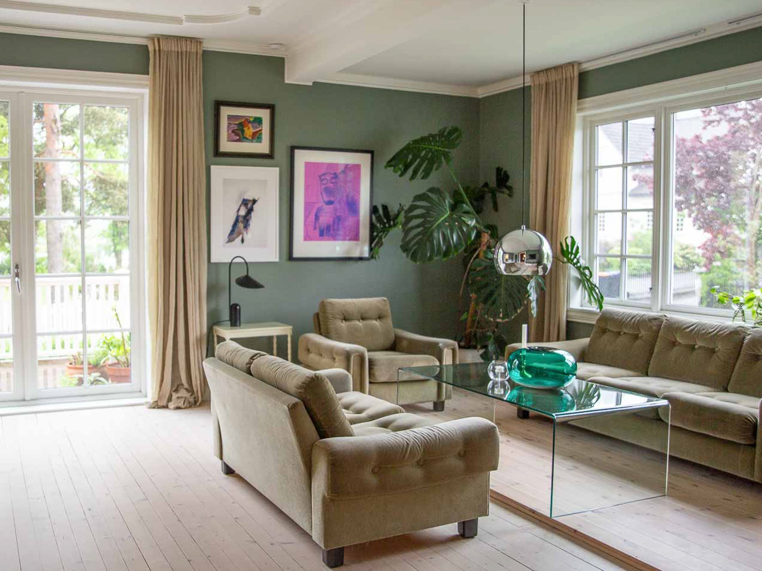 Grønn stue malt i Jotun Antique green med vintage møbler, fargerik kunst og stor grønn plante i hjørnet.