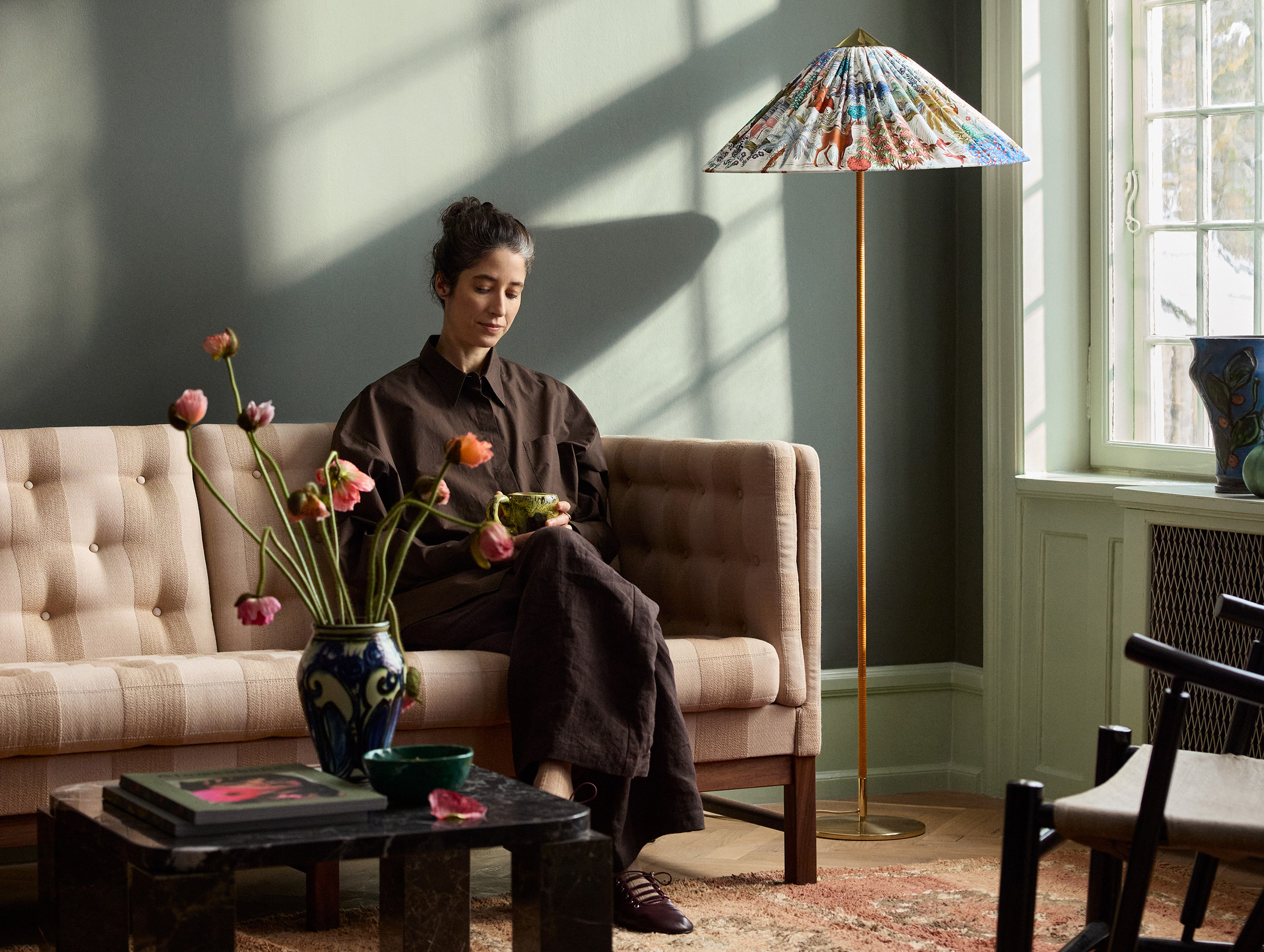 En kvinna sitter på en beige soffa, håller i en kopp, i ett solbelyst vardagsrum med en färgglad golvlampa och en blomvas på ett soffbord. Vägg målad i Jotun 7613 Northern Mystic.