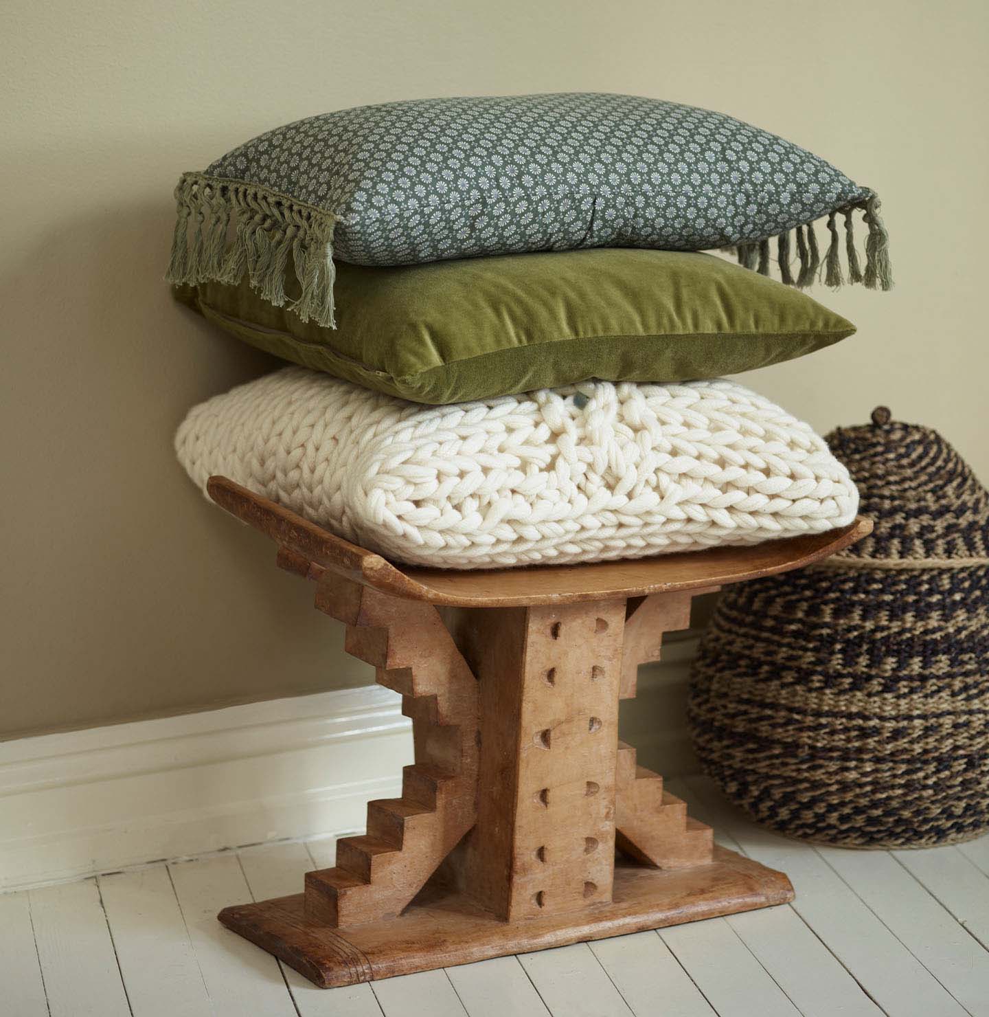 Stack of textured cushions on a carved wooden stool, with a woven basket beside it, set against a muted, beige yellow wall painted with Jotun 1938 Tea Leaves.