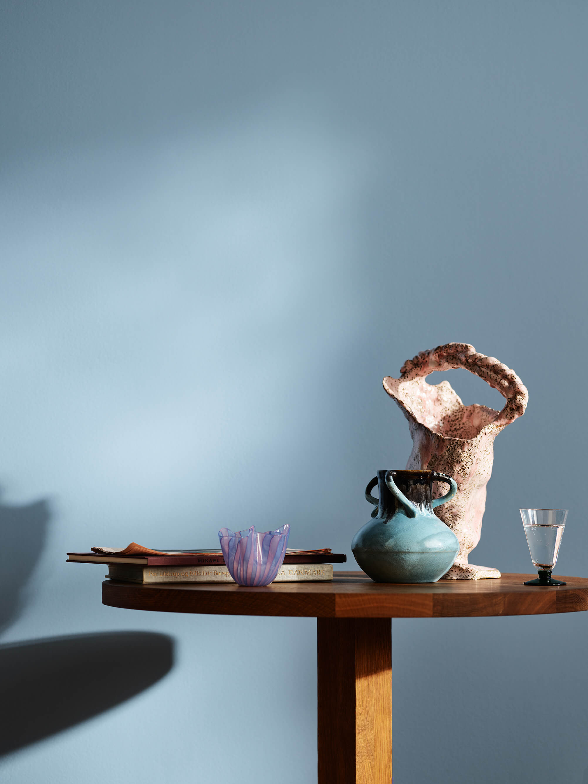 Kjøkken med blå vegger i fargen Jotun 4223 Sama. På et trebord står det bøker, en blå keramikkkrukke, en rosa vase med struktur og et lite glass.