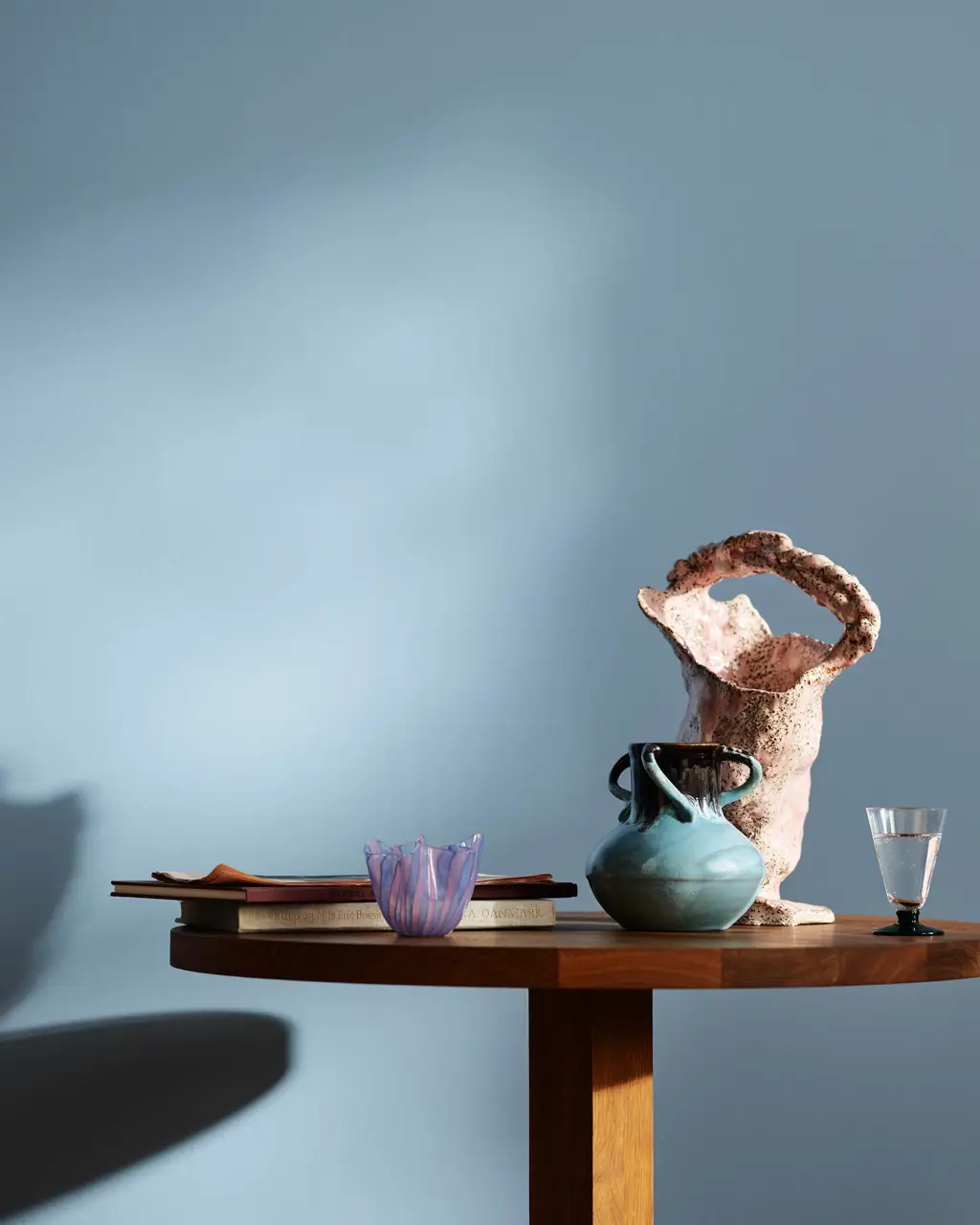 Kjøkken med blå vegger i fargen Jotun 4223 Sama. På et trebord står det bøker, en blå keramikkkrukke, en rosa vase med struktur og et lite glass.