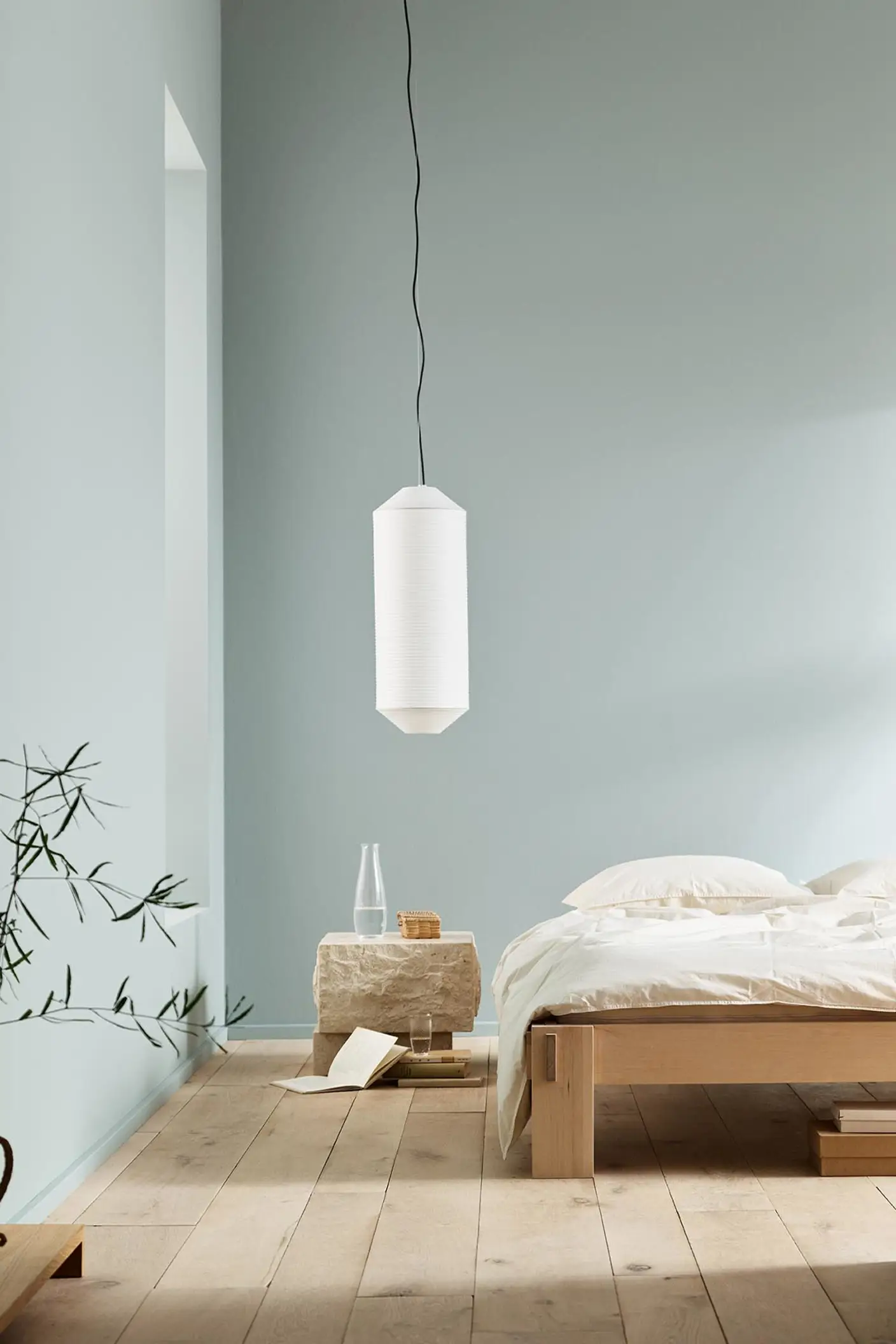 Minimalist bedroom with white bedding, a stone side table, and a plant against muted blue walls painted with the colour 6378 Iconic from Jotun.