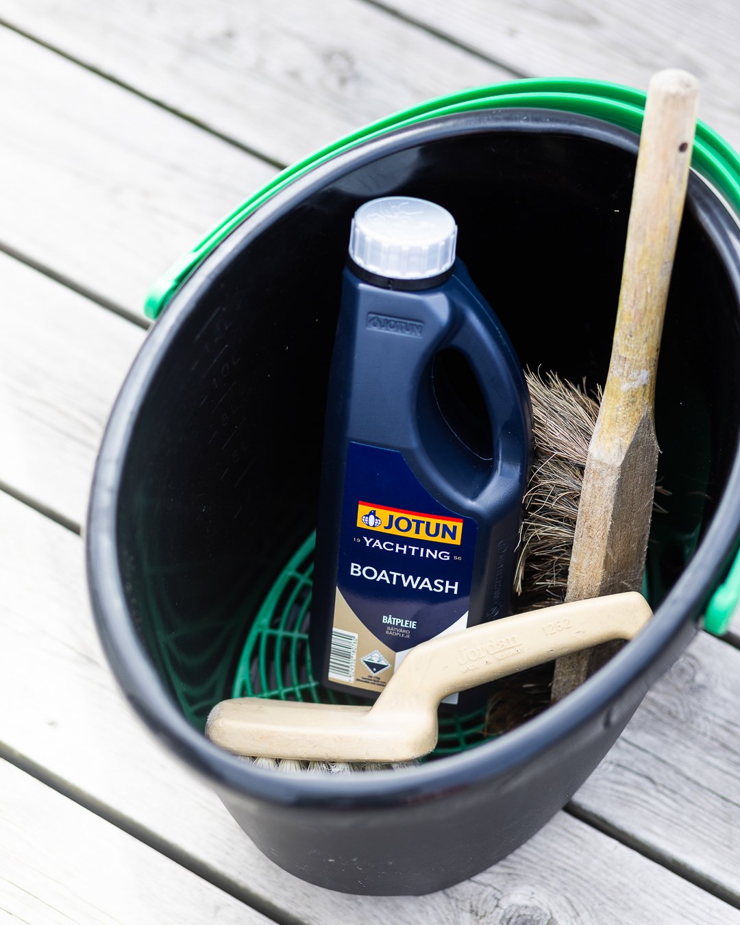 Emballasje med Jotun Boatwash, kost og børste i en bøtte på brygge med vann i bakgrunnen.
