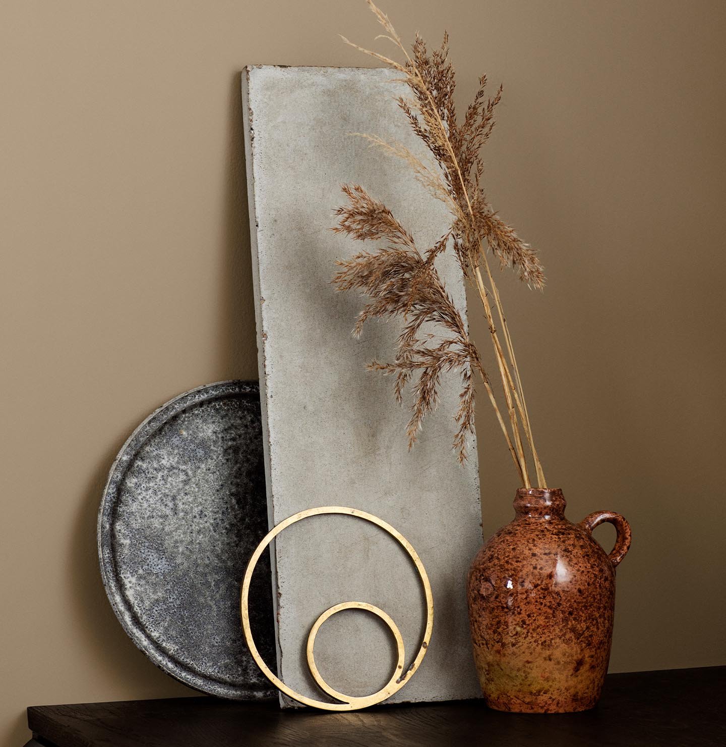 Keramikkanna, guldringar och betongskiva mot en brun, gyllene vägg målad med färgen 10962 Sandy från Jotun.