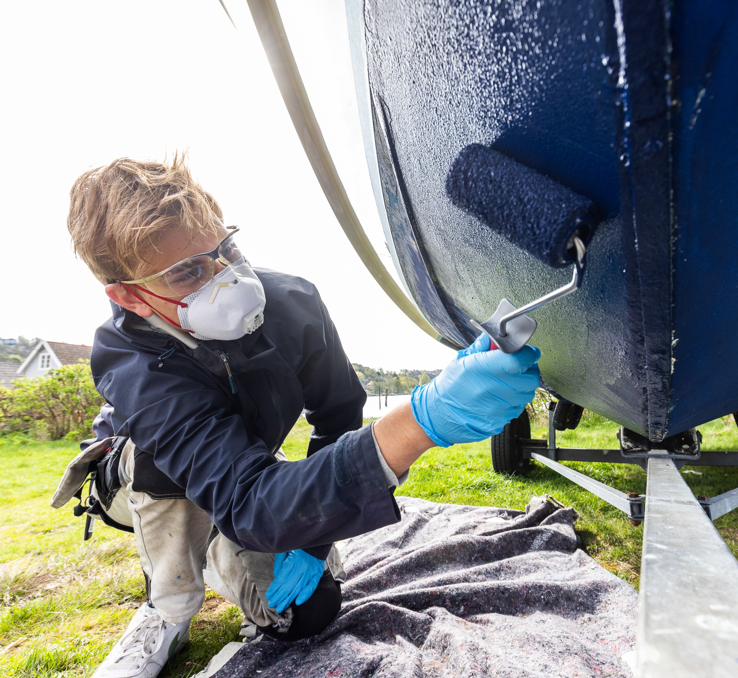 Ung mann med maske og beskyttelsesbriller ruller blått bunnstoff på undersiden av en fritidsbåt