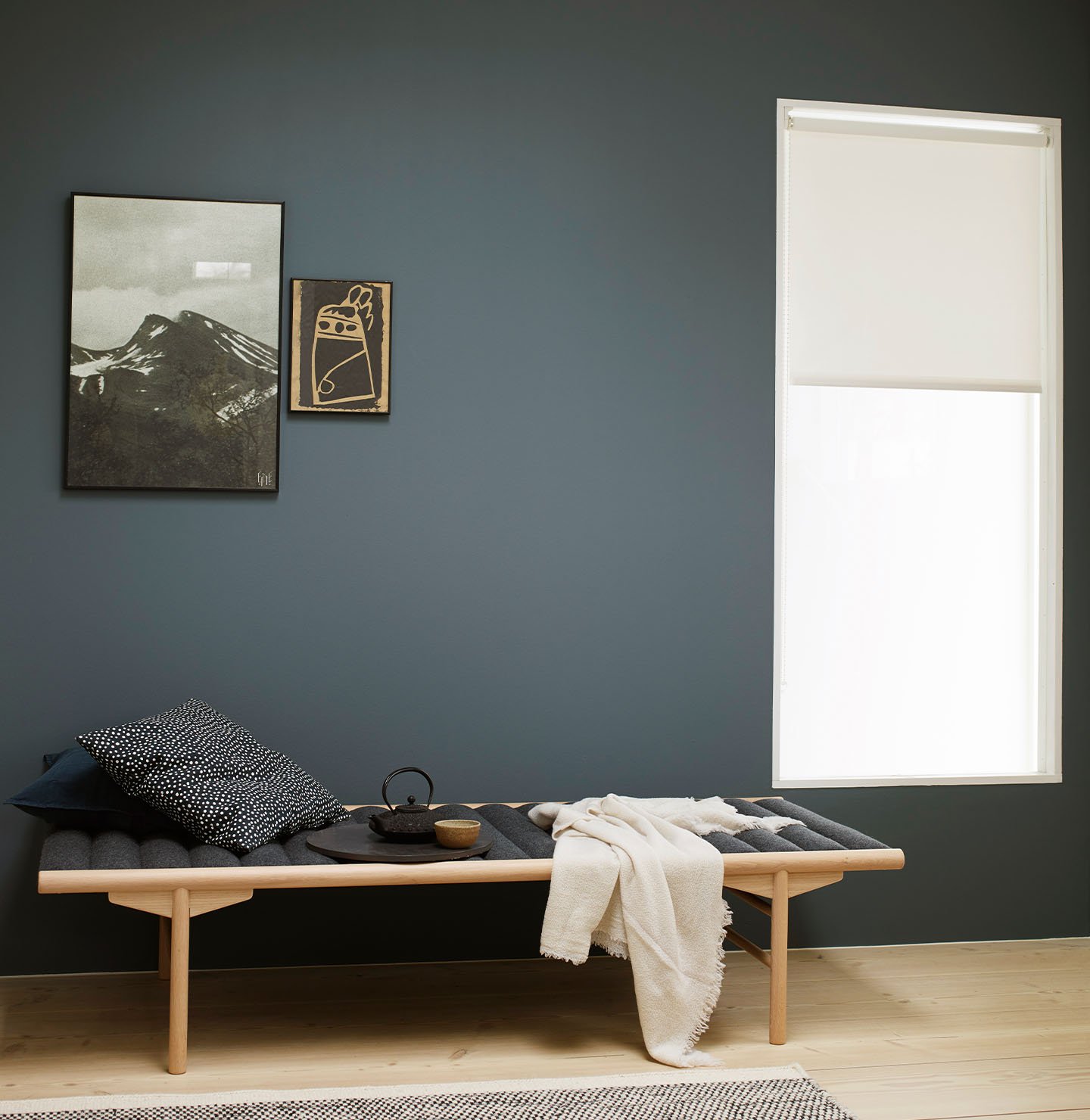 Ett rogivande utrymme med en träbänk lagerlagd med mörka kuddar och en krämfärgad pläd, placerad mot en dämpad blågrön vägg målad med Jotun 5030 St. Paul Blue. Taket är målat i Jotun 5249 Linblå, en mjukt blågrön nyans.