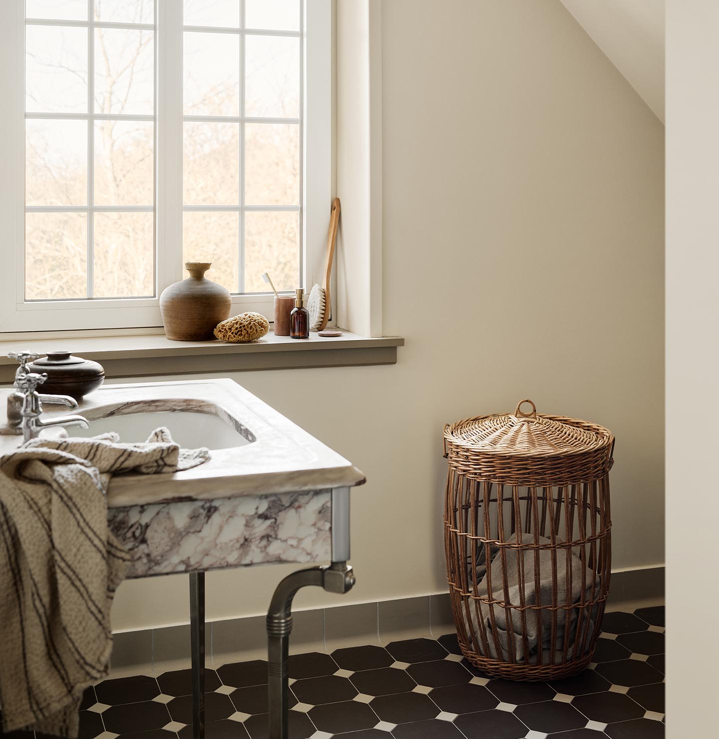 Bathroom with a marble sink, patterned floor tiles, and a wicker laundry basket against a wall painted with 12079 Gleam - a muted, greenish yellow colour from Jotun.