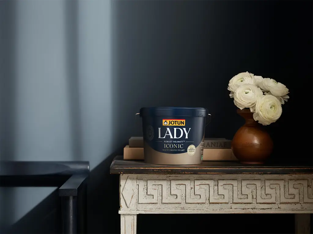 JOTUN LADY Iconic färgburk på ett bord med vita blommor i vas mot mörk vägg