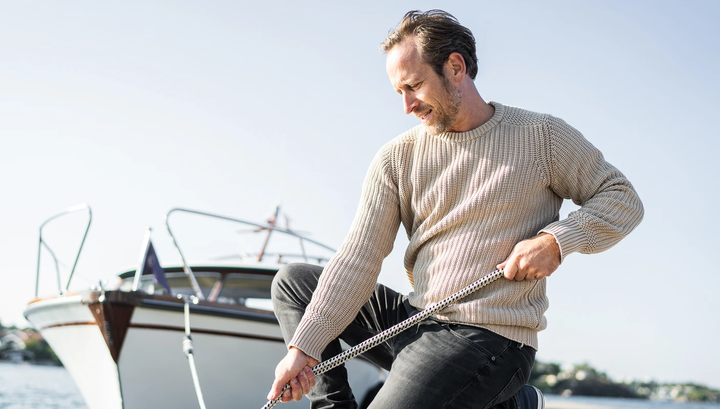 Mann drar i tau på bryggekant med båt og blå himmel i bakgrunnen