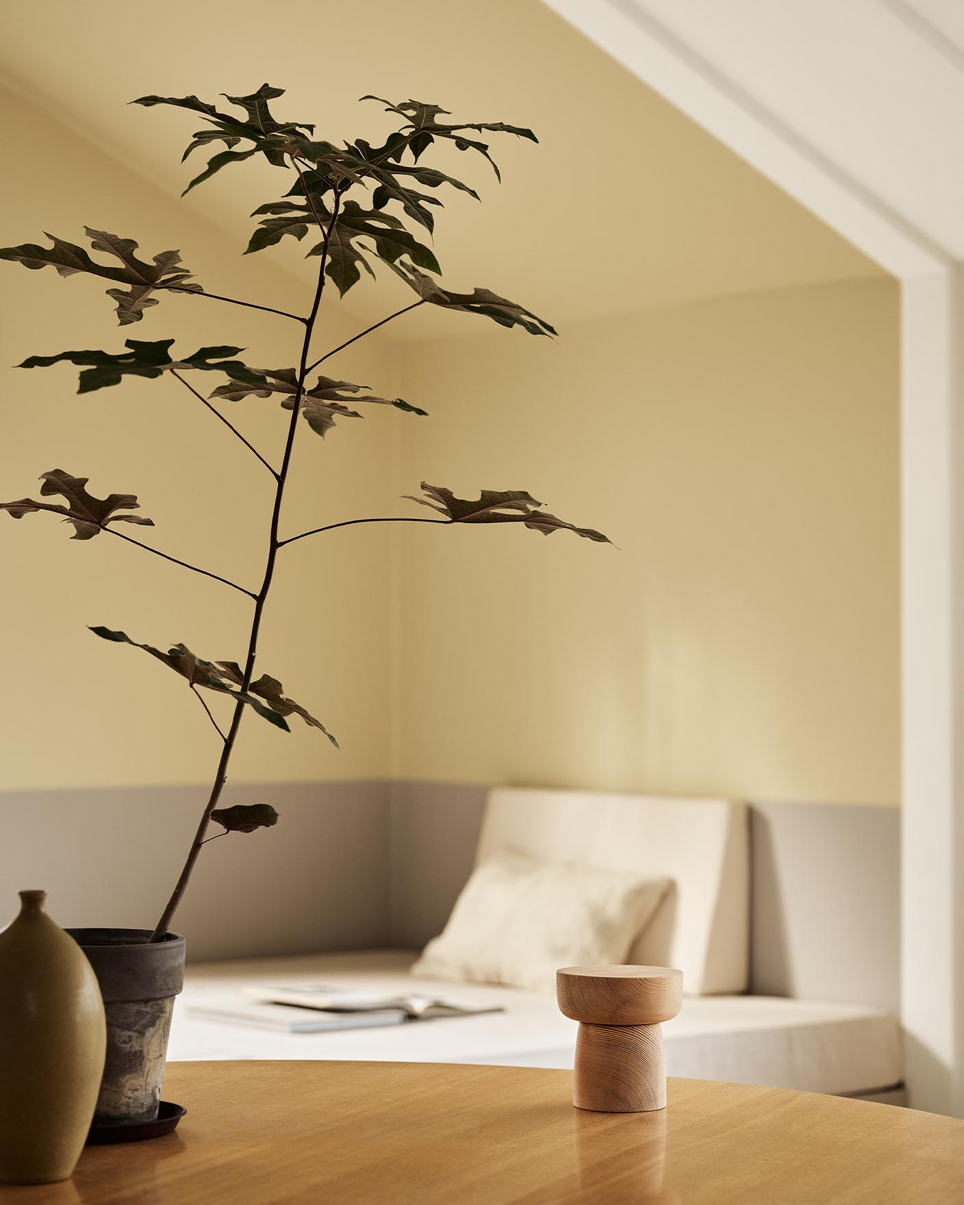 A potted plant and a daybed with light bedding against muted, greenish yellow walls painted in the colour 1965 Ginger Tea from Jotun.