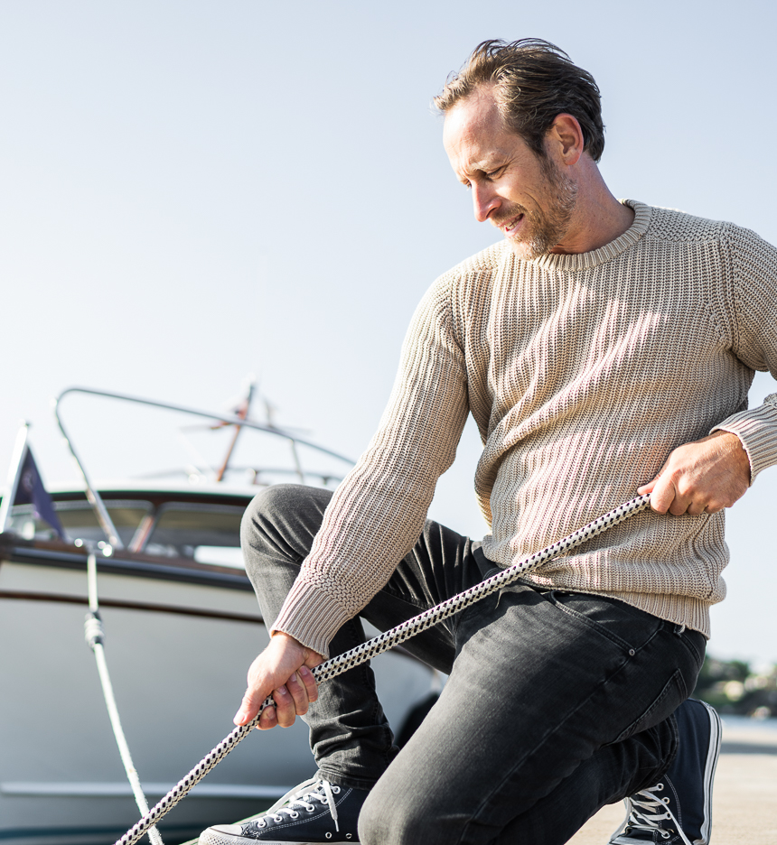 Mann drar i tau på bryggekant med båt og blå himmel i bakgrunnen.