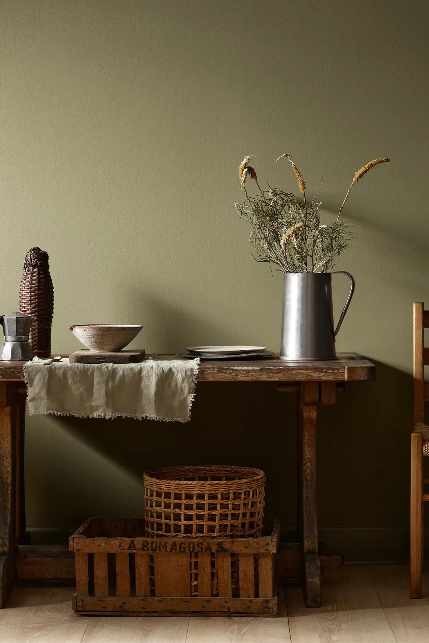 Rustic wooden table with vintage kitchenware and a wicker basket against a olivegreen wall painted with the colour 8597 Seaweed Green from Jotun.