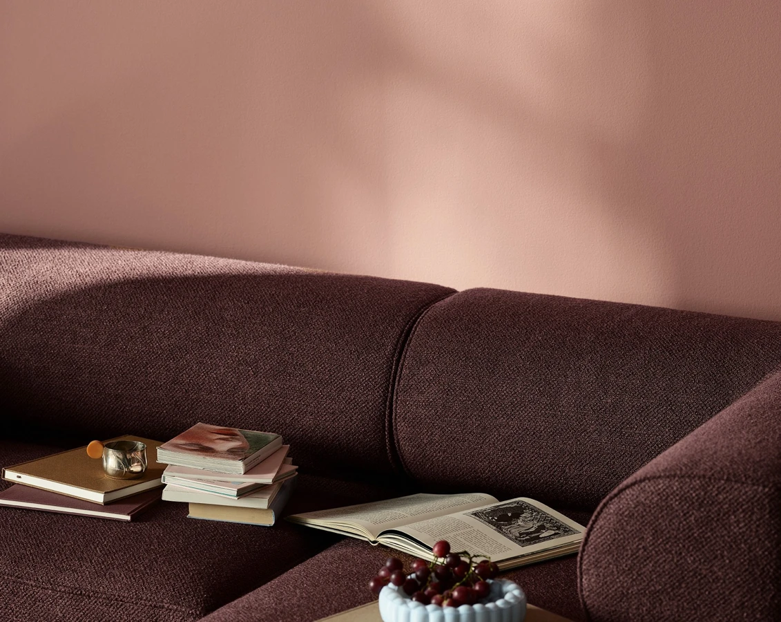En stue med vegger malt i Jotun 20256 Balmy Pink, med en plommefarget sofa med bøker og en bolle med druer.