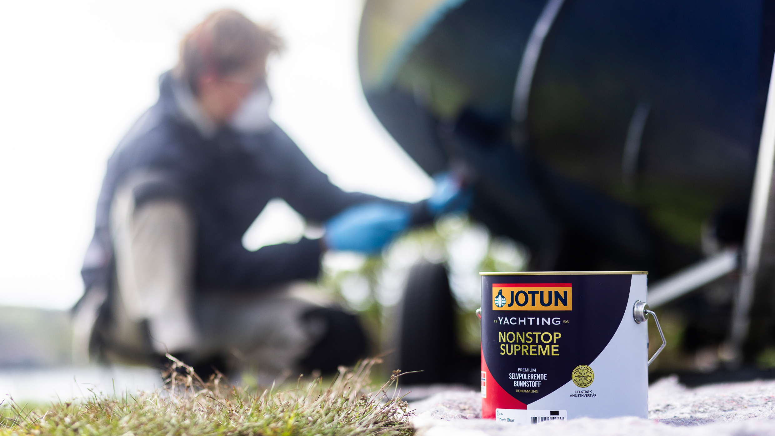 Mann med beskyttelsesmaske og briller legger blått bunnstoff på båt, i front sees et spann fra Jotun Yachting med bunnstoffet NonStop Supreme