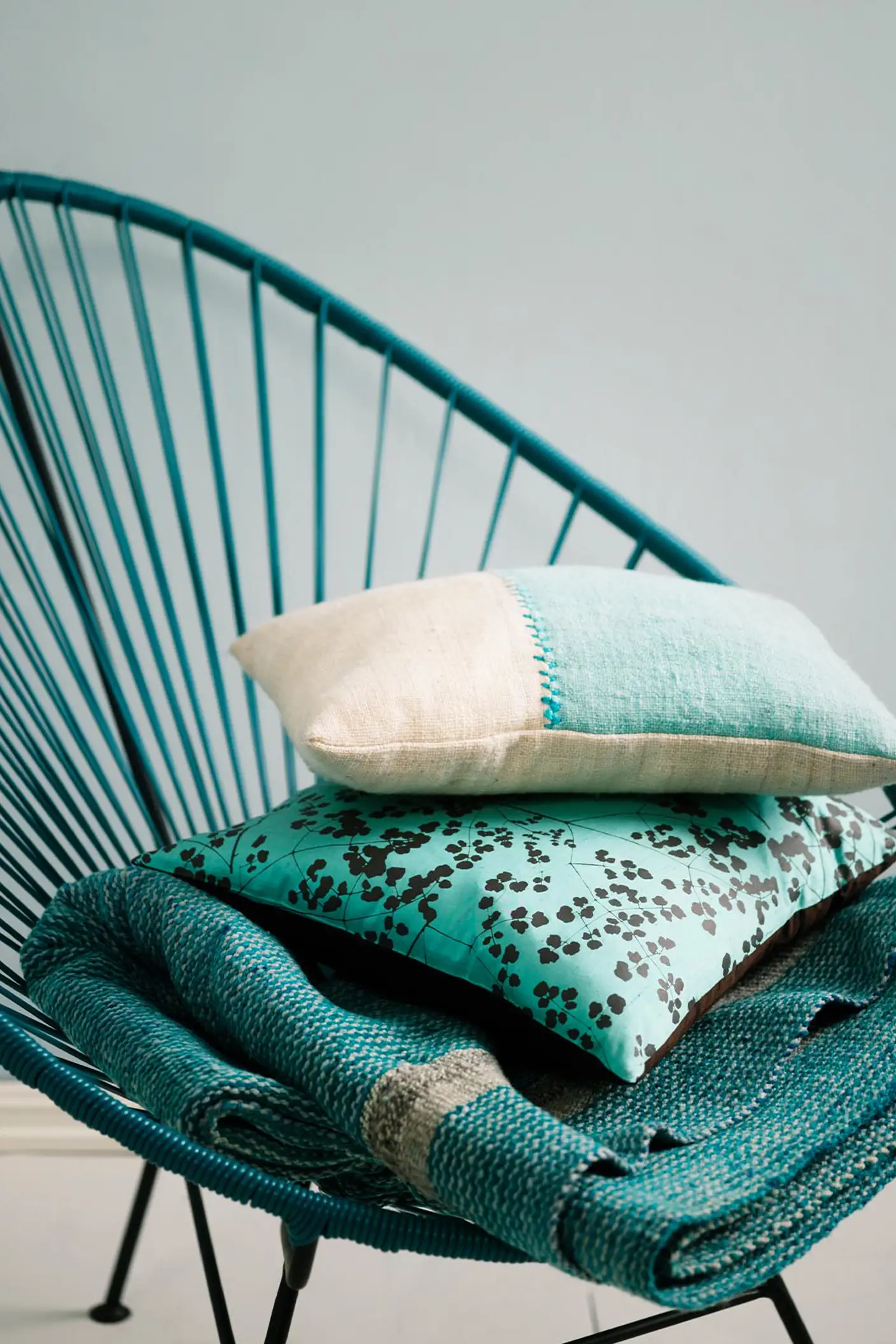 Teal woven chair holding a stack of textured cushions and a blanket in turquoise and cream tones, set against a muted, greenish blue wall painted with Jotun 5225 Lake View.