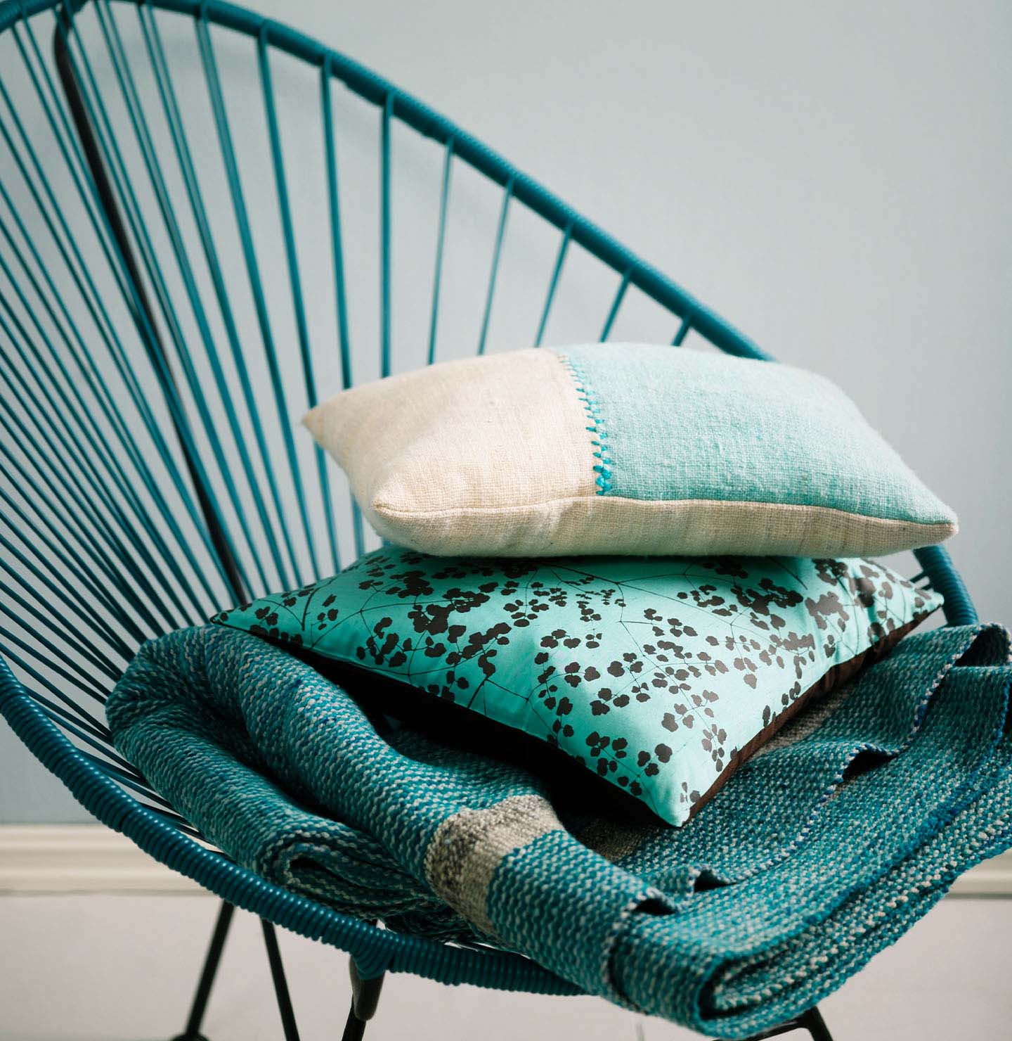 Teal woven chair holding a stack of textured cushions and a blanket in turquoise and cream tones, set against a muted, greenish blue wall painted with Jotun 5225 Lake View.