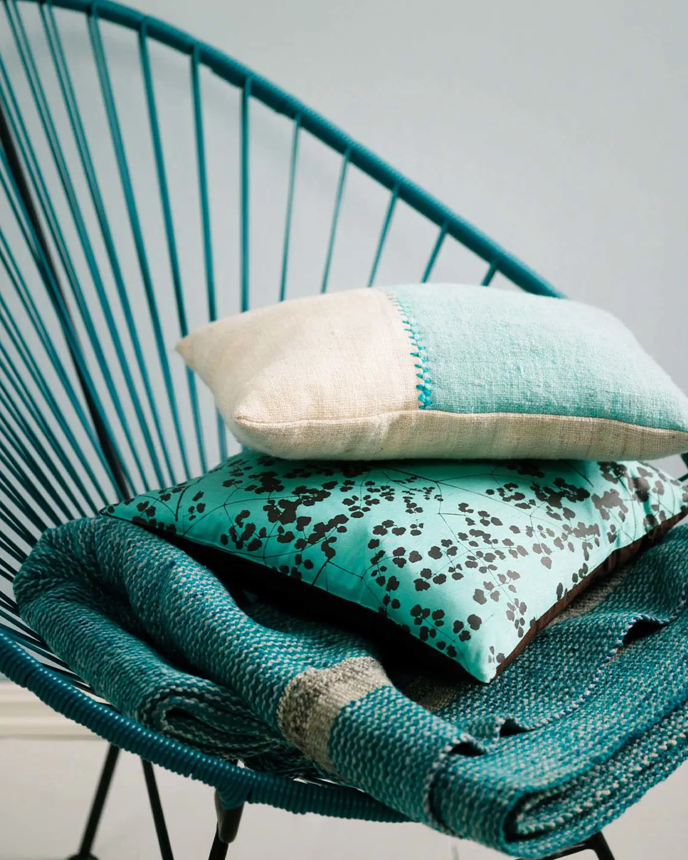 Teal woven chair holding a stack of textured cushions and a blanket in turquoise and cream tones, set against a muted, greenish blue wall painted with Jotun 5225 Lake View.