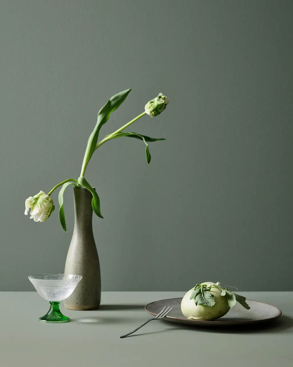 En tulpan i en vas på ett bord i mjuka gröna toner målat med Jotun 7685 Subtle Green, med en bakgrundsvägg målad i 7686 Mindful Green från Jotun.