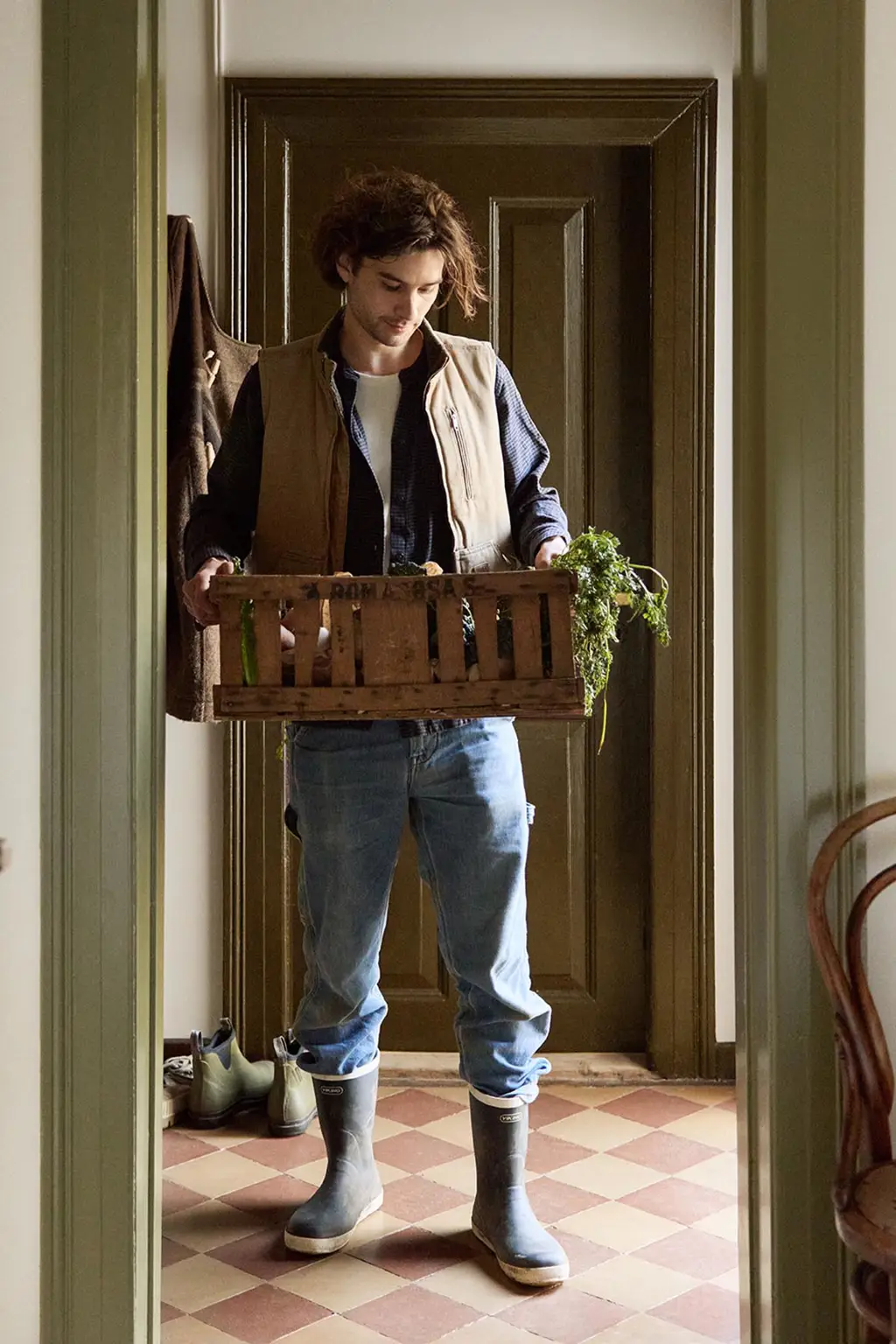 Person in a doorway holding a crate with greenery. Walls painted in the muted yellow colour 12079 Gleam from Jotun. Olive-green trims in Jotun's 8597 Seawood Green.