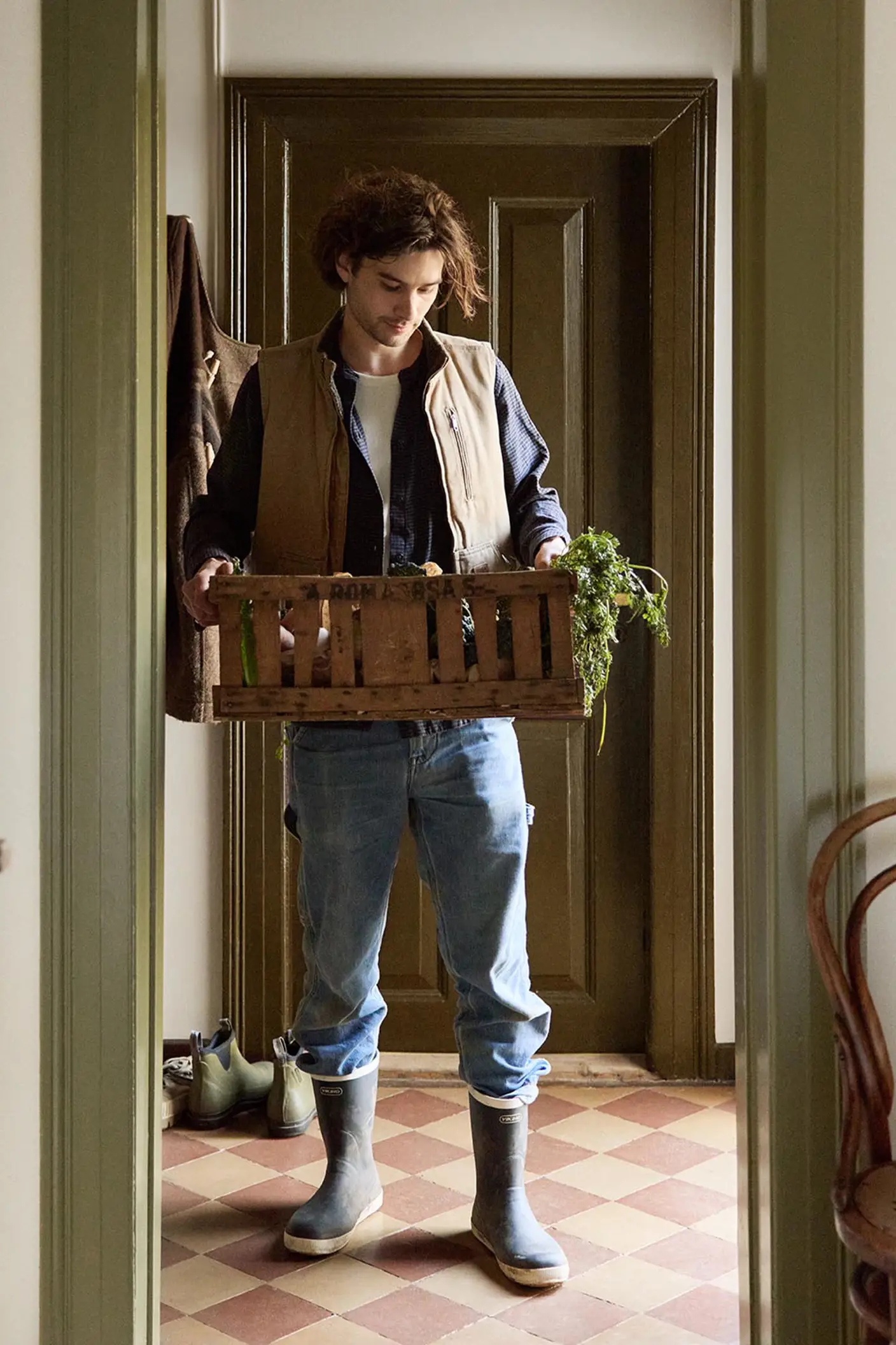 Person in a doorway holding a crate with greenery. Walls painted in the muted yellow colour 12079 Gleam from Jotun. Olive-green trims in Jotun's 8597 Seawood Green.
