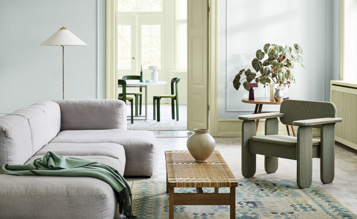 Modern living room with gray sofa, green armchair, woven coffee table, and potted plants, leading to a bright dining area with black chairs