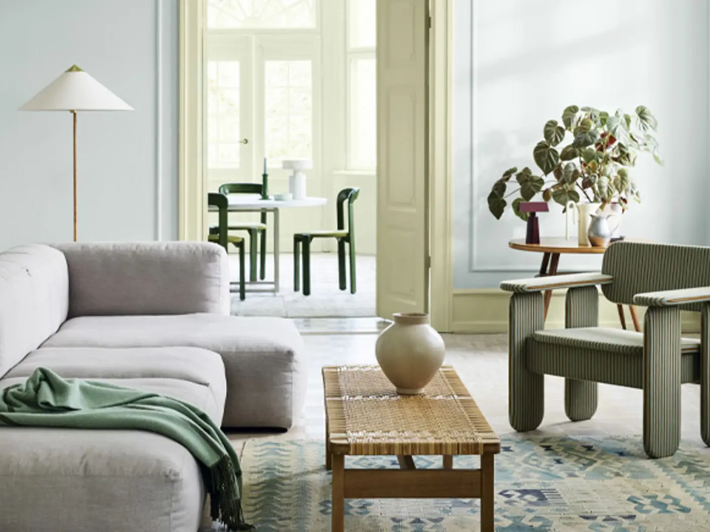 Modern living room with gray sofa, green armchair, woven coffee table, and potted plants, leading to a bright dining area with black chairs