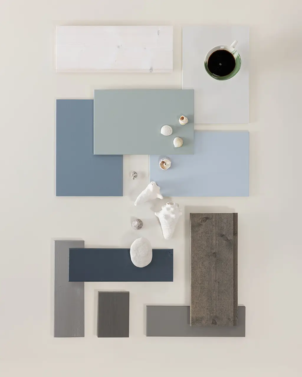 Interiördesigninspirerad flatlay med blå och grå färgtoner, olika texturer av färgprover, träslag och små snäckor, samt en kopp kaffe.
