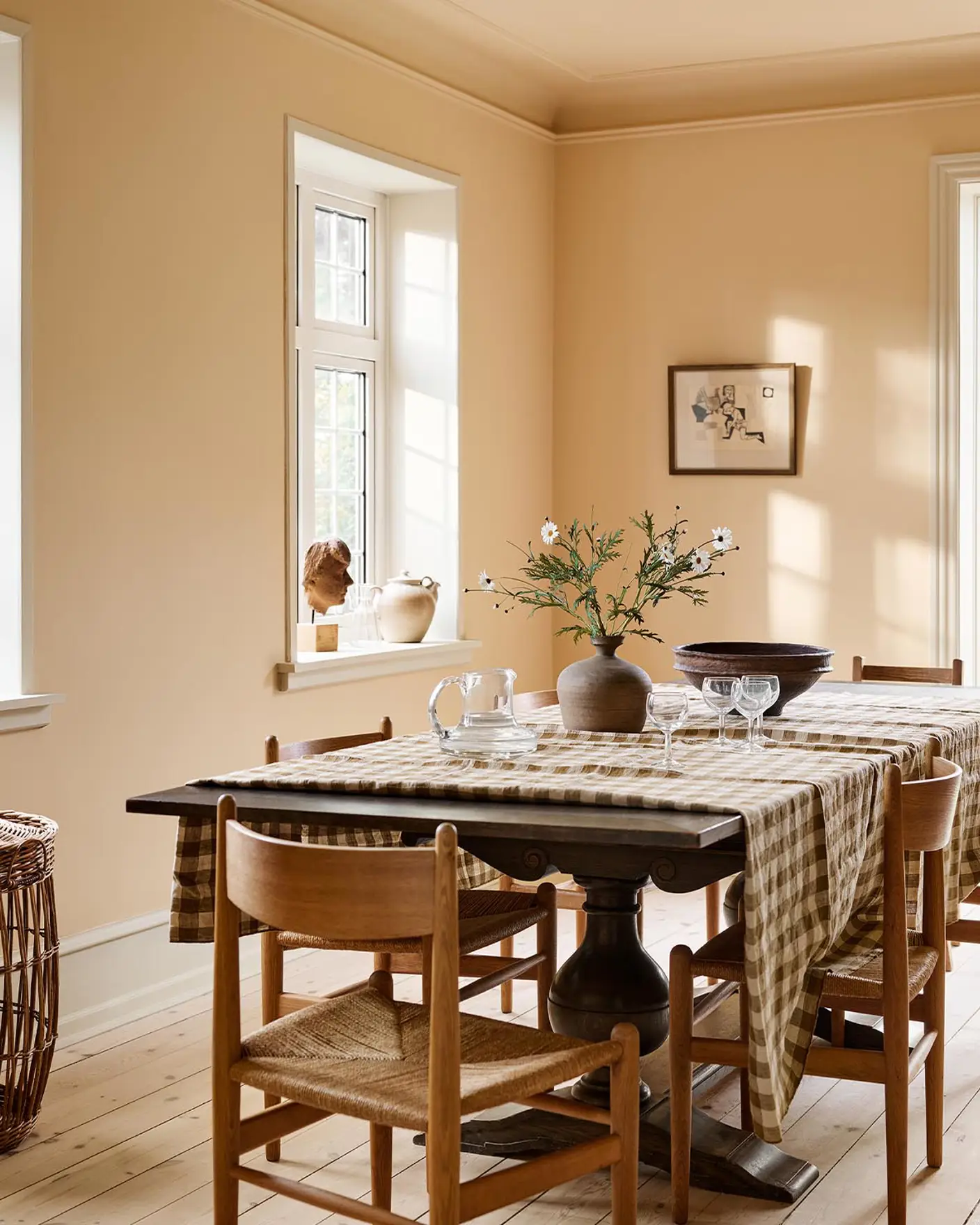 Dining room with a checkered tablecloth, wooden chairs, against an ochre yellow wall painted in the colour 11202 Mild Ochre from Jotun.